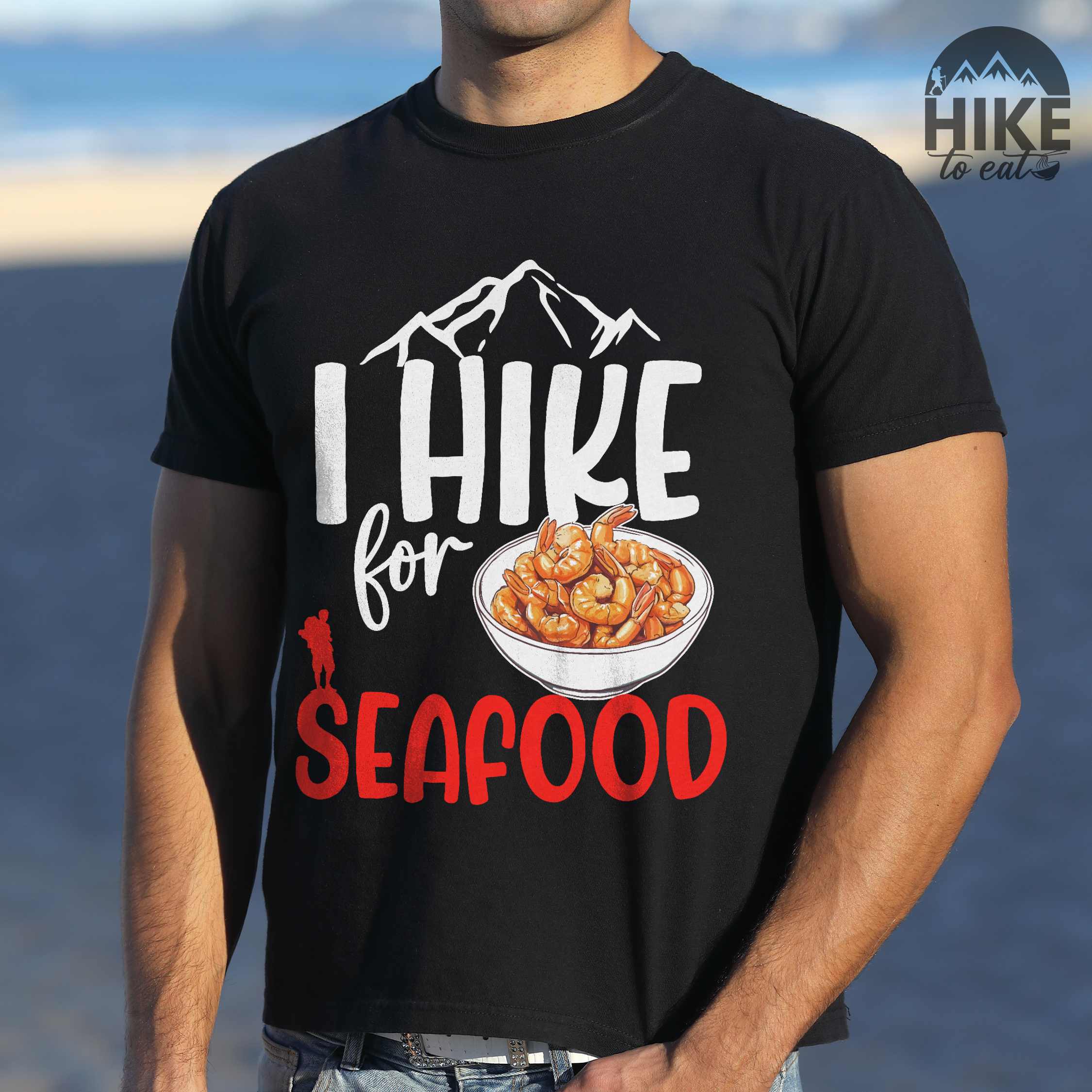 Man wearing black "I hike for seafood" colored vintage tee with bold typography, illustrated shrimps on a bowl, mountains, birds, and backpackers for seafood lovers.