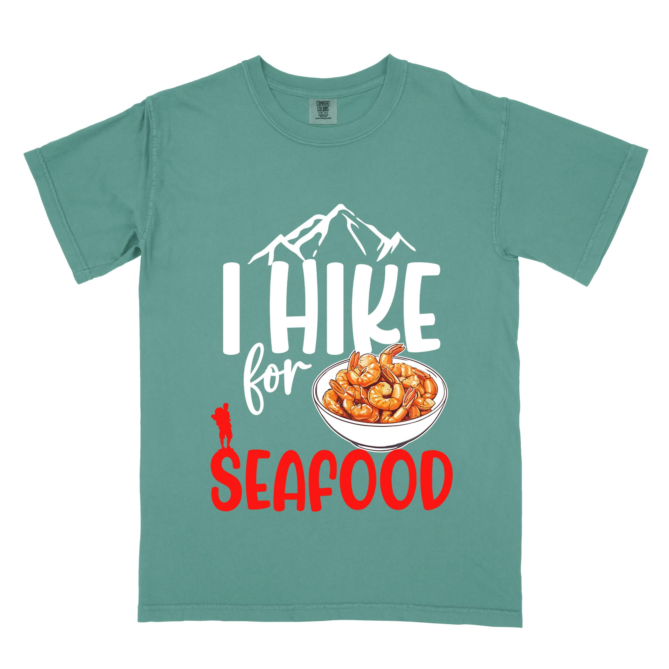 Light green "I hike for seafood" colored vintage tee with bold typography, illustrated shrimps on a bowl, mountains, birds, and backpackers for seafood lovers.
