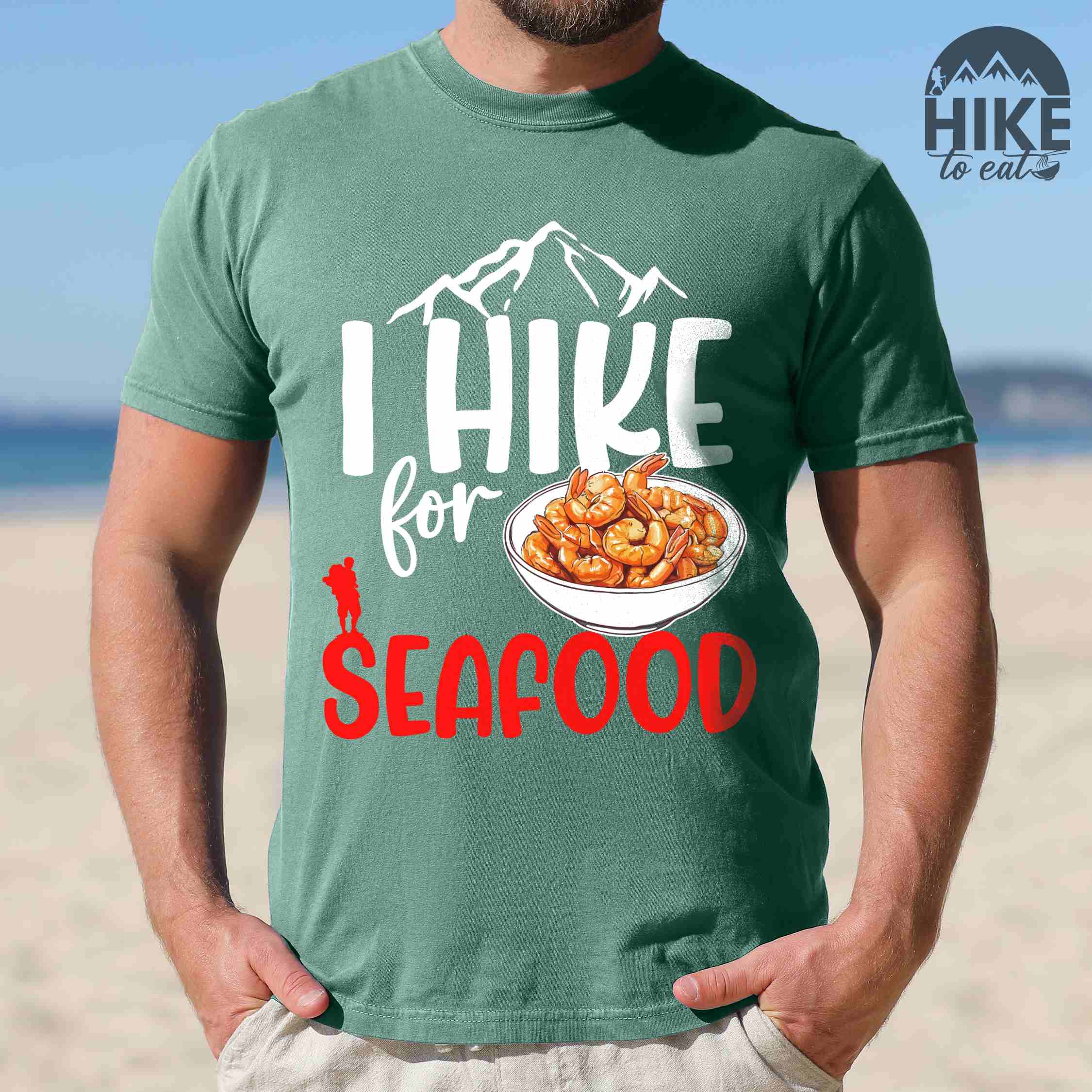 Man wearing light green "I hike for seafood" colored vintage tee with bold typography, illustrated shrimps on a bowl, mountains, birds, and backpackers for seafood lovers.