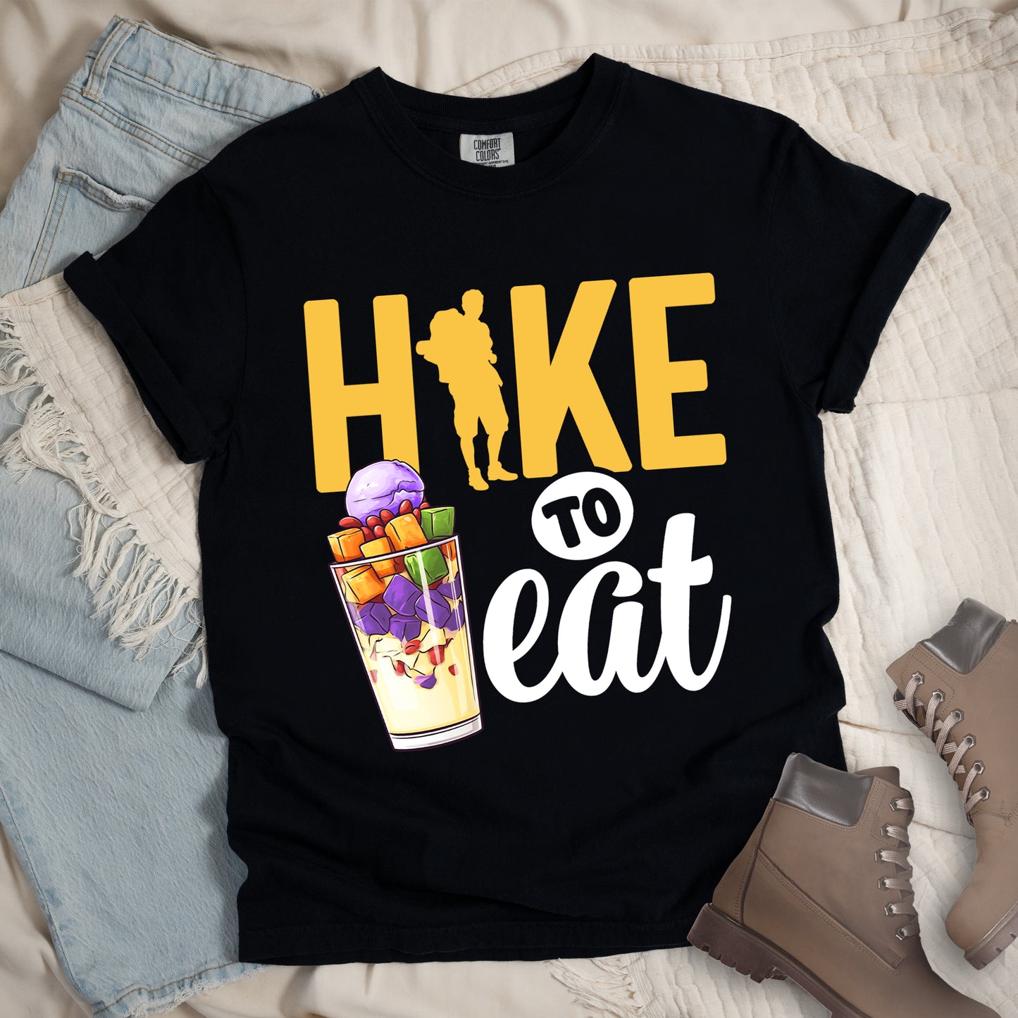 Black t-shirt with yellow and white text reading "HIKE TO eat". "HIKE" appears in large yellow letters at the top with a yellow hiker silhouette integrated into the letter "I", "TO" in black text on a white circular background in the middle, and "eat" in white script at the bottom. Features an illustration of a colorful fruit parfait/dessert (Halo-Halo) in a clear glass with purple, orange, and green fruit pieces to the left of "TO eat".
