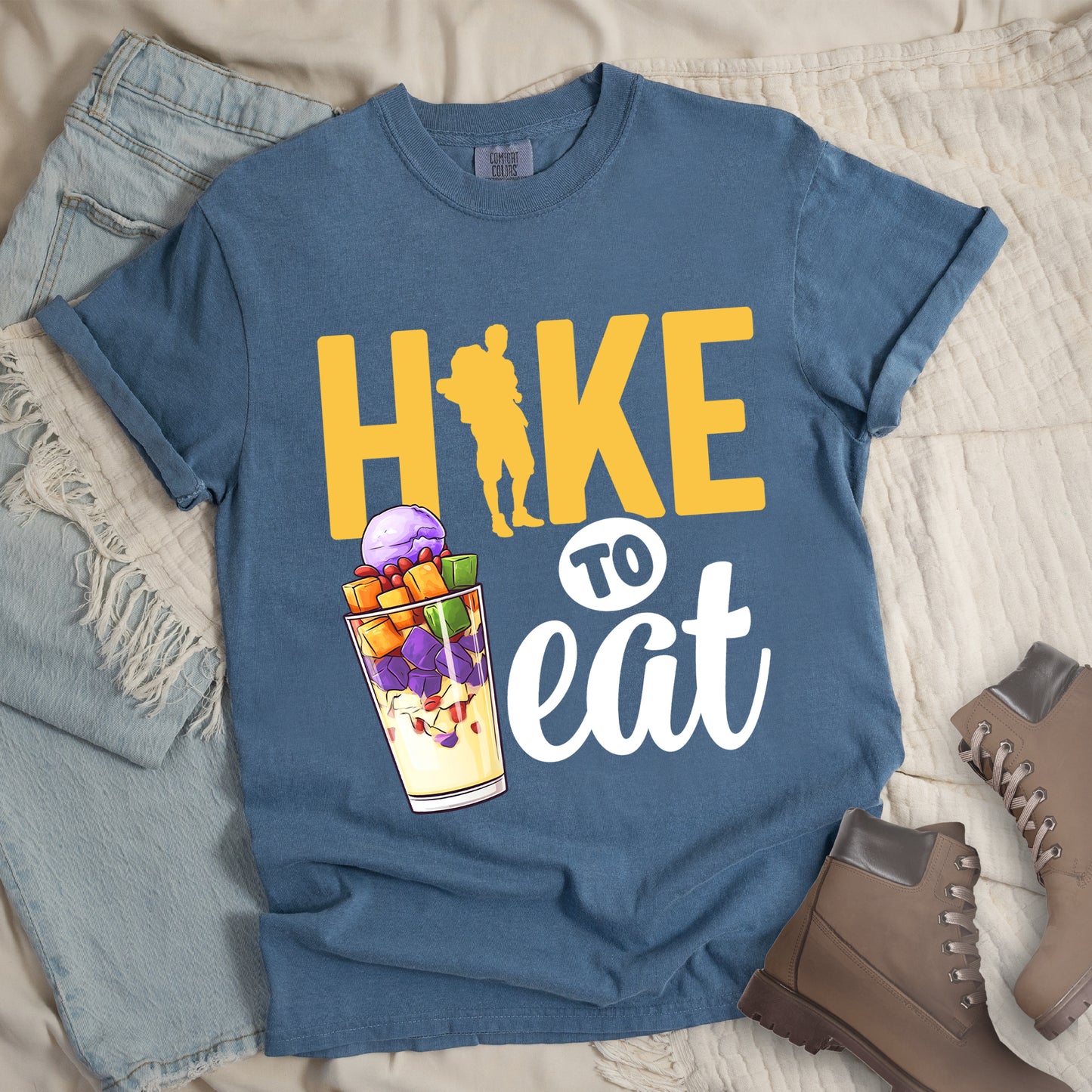 Blue Jean (Denim Blue) t-shirt with yellow and white text reading "HIKE TO eat". "HIKE" appears in large yellow letters at the top with a yellow hiker silhouette integrated into the letter "I", "TO" in black text on a white circular background in the middle, and "eat" in white script at the bottom. Features an illustration of a colorful fruit parfait/dessert (Halo-Halo) in a clear glass with purple, orange, and green fruit pieces to the left of "TO eat".