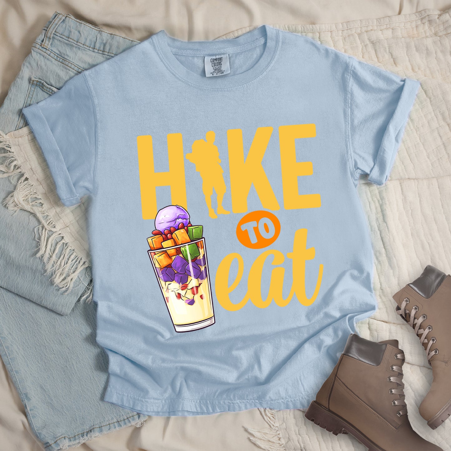 Chambray (Light Blue) t-shirt with yellow text reading "HIKE TO eat". "HIKE" appears in large yellow letters at the top with a yellow hiker silhouette integrated into the letter "I", "TO" in Chambray (Light Blue) text on an orange circular background in the middle, and "eat" in yellow script at the bottom. Features an illustration of a colorful fruit parfait/dessert (Halo-Halo) in a clear glass with purple, orange, and green fruit pieces to the left of "TO eat".