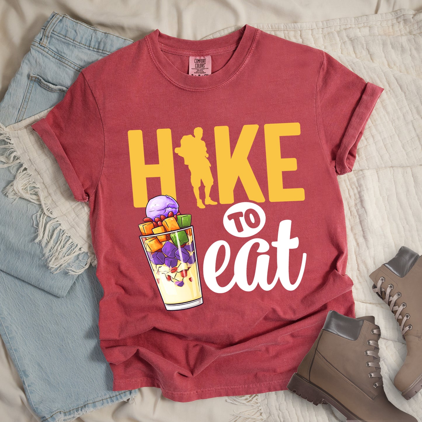 Crimson (Dark Pink) t-shirt with yellow and white text reading "HIKE TO eat". "HIKE" appears in large yellow letters at the top with a yellow hiker silhouette integrated into the letter "I", "TO" in black text on a white circular background in the middle, and "eat" in white script at the bottom. Features an illustration of a colorful fruit parfait/dessert (Halo-Halo) in a clear glass with purple, orange, and green fruit pieces to the left of "TO eat".