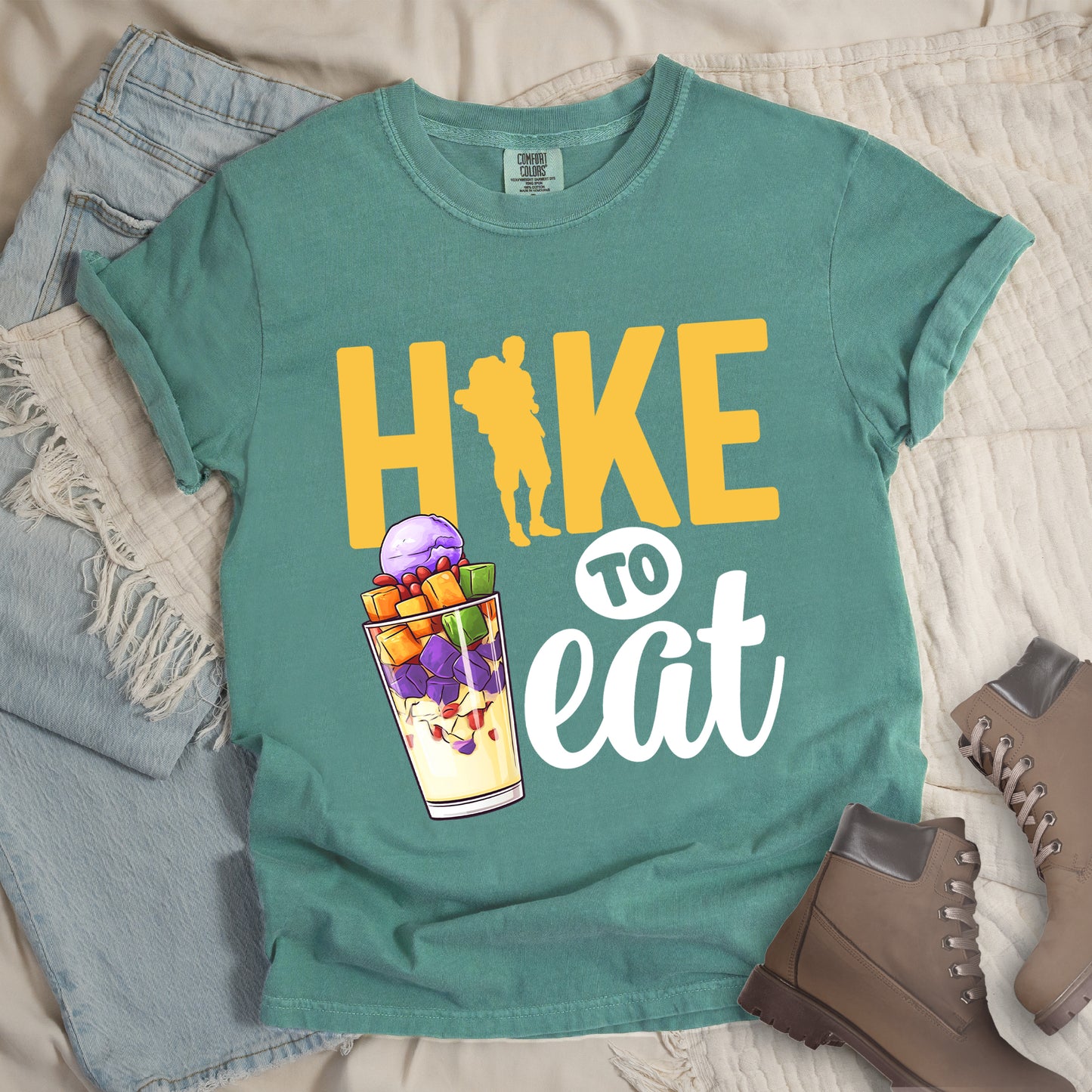 Light Green t-shirt with yellow and white text reading "HIKE TO eat". "HIKE" appears in large yellow letters at the top with a yellow hiker silhouette integrated into the letter "I", "TO" in black text on a white circular background in the middle, and "eat" in white script at the bottom. Features an illustration of a colorful fruit parfait/dessert (Halo-Halo) in a clear glass with purple, orange, and green fruit pieces to the left of "TO eat".