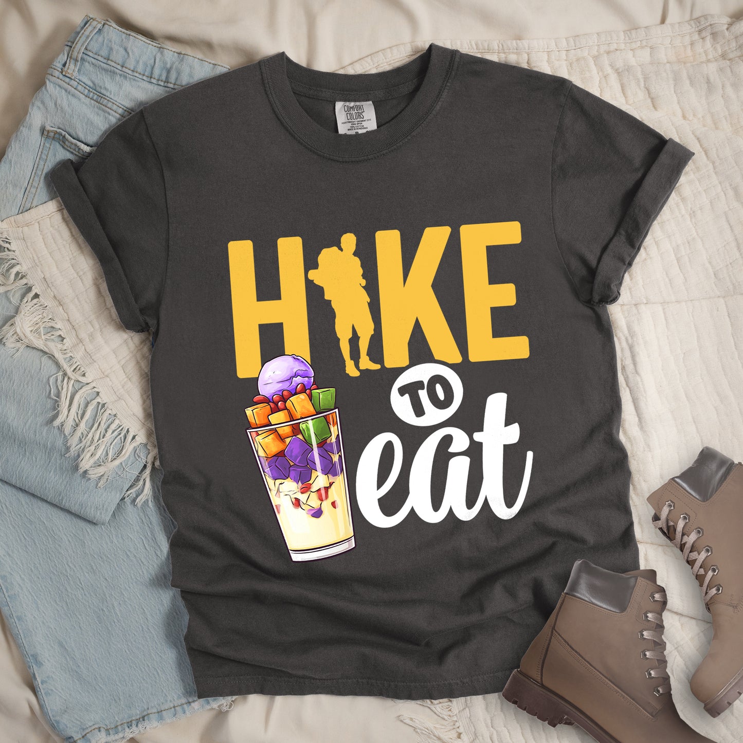 Pepper (Dark Gray) t-shirt with yellow and white text reading "HIKE TO eat". "HIKE" appears in large yellow letters at the top with a yellow hiker silhouette integrated into the letter "I", "TO" in black text on a white circular background in the middle, and "eat" in white script at the bottom. Features an illustration of a colorful fruit parfait/dessert (Halo-Halo) in a clear glass with purple, orange, and green fruit pieces to the left of "TO eat".