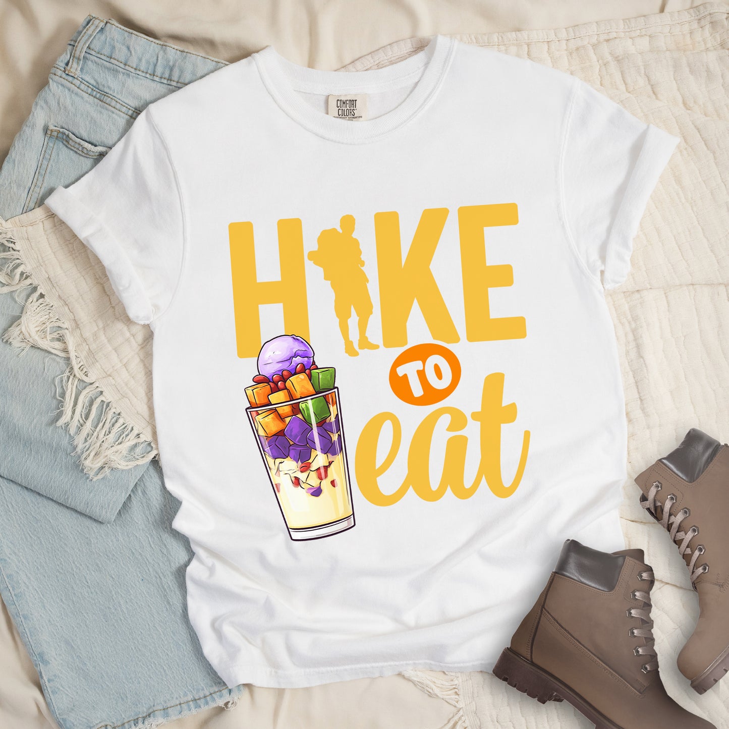 White t-shirt with yellow text reading "HIKE TO eat". "HIKE" appears in large yellow letters at the top with a yellow hiker silhouette integrated into the letter "I", "TO" in white text on an orange circular background in the middle, and "eat" in yellow script at the bottom. Features an illustration of a colorful fruit parfait/dessert (Halo-Halo) in a clear glass with purple, orange, and green fruit pieces to the left of "TO eat".