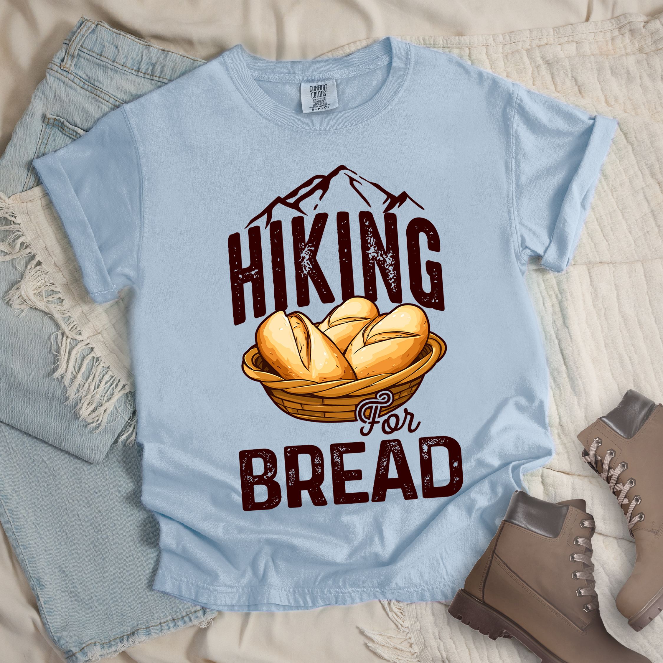 Light blue t-shirt with brown distressed text reading "HIKING For BREAD". "HIKING" appears in large brown distressed letters at the top with brown mountain outlines above it, "For" in small brown script in the middle, and "BREAD" in large brown distressed letters at the bottom. Features an illustration of bread rolls in a woven basket between the words "HIKING" and "BREAD". Features a classic crew neck and casual rolled sleeves, styled alongside light wash denim and brown hiking boots.