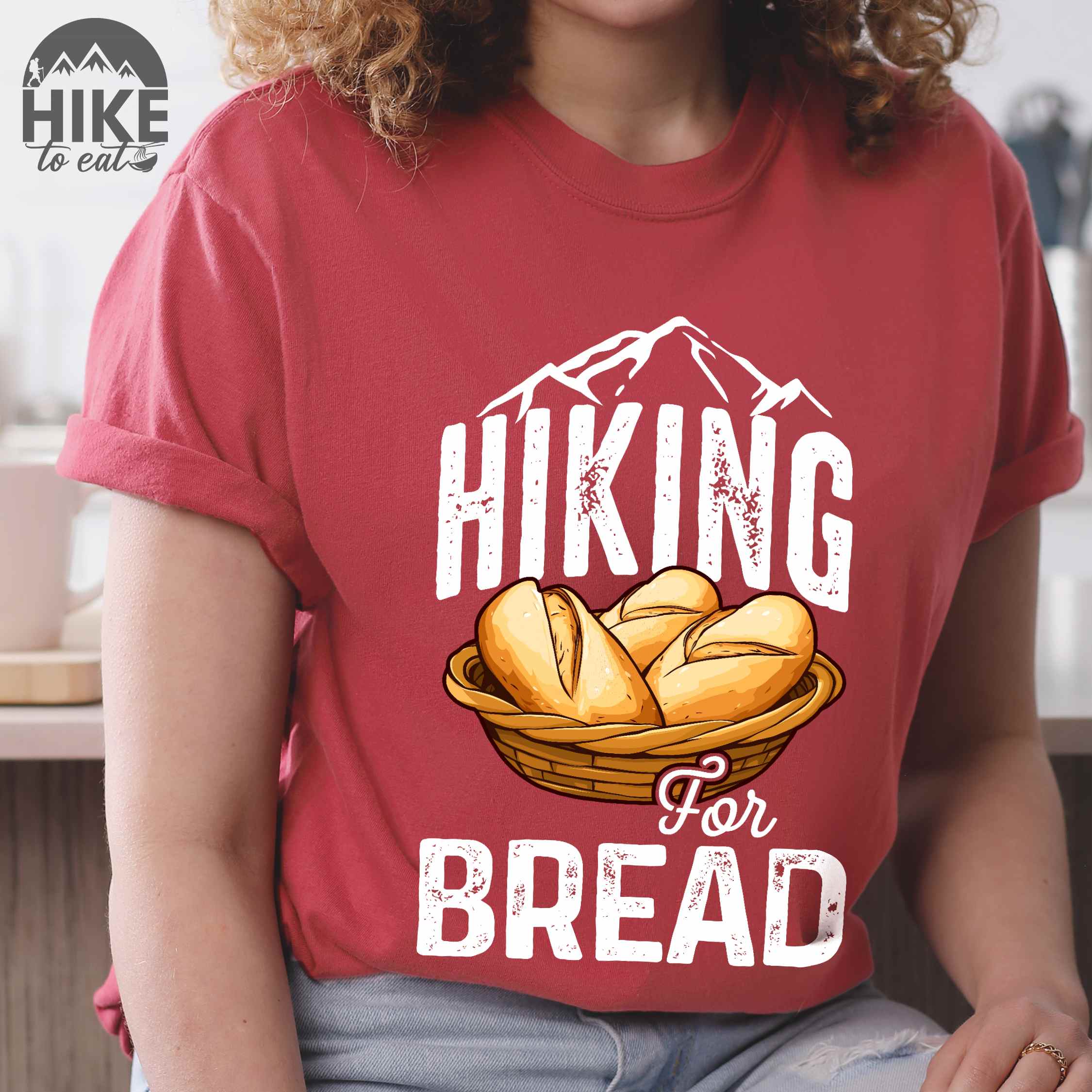 Woman wearing crimson "Hiking for bread" colored statement tee with flowing script typography, bold lettering, and freshly baked bread illustration for pastry-loving outdoor enthusiasts, hikers and foodies