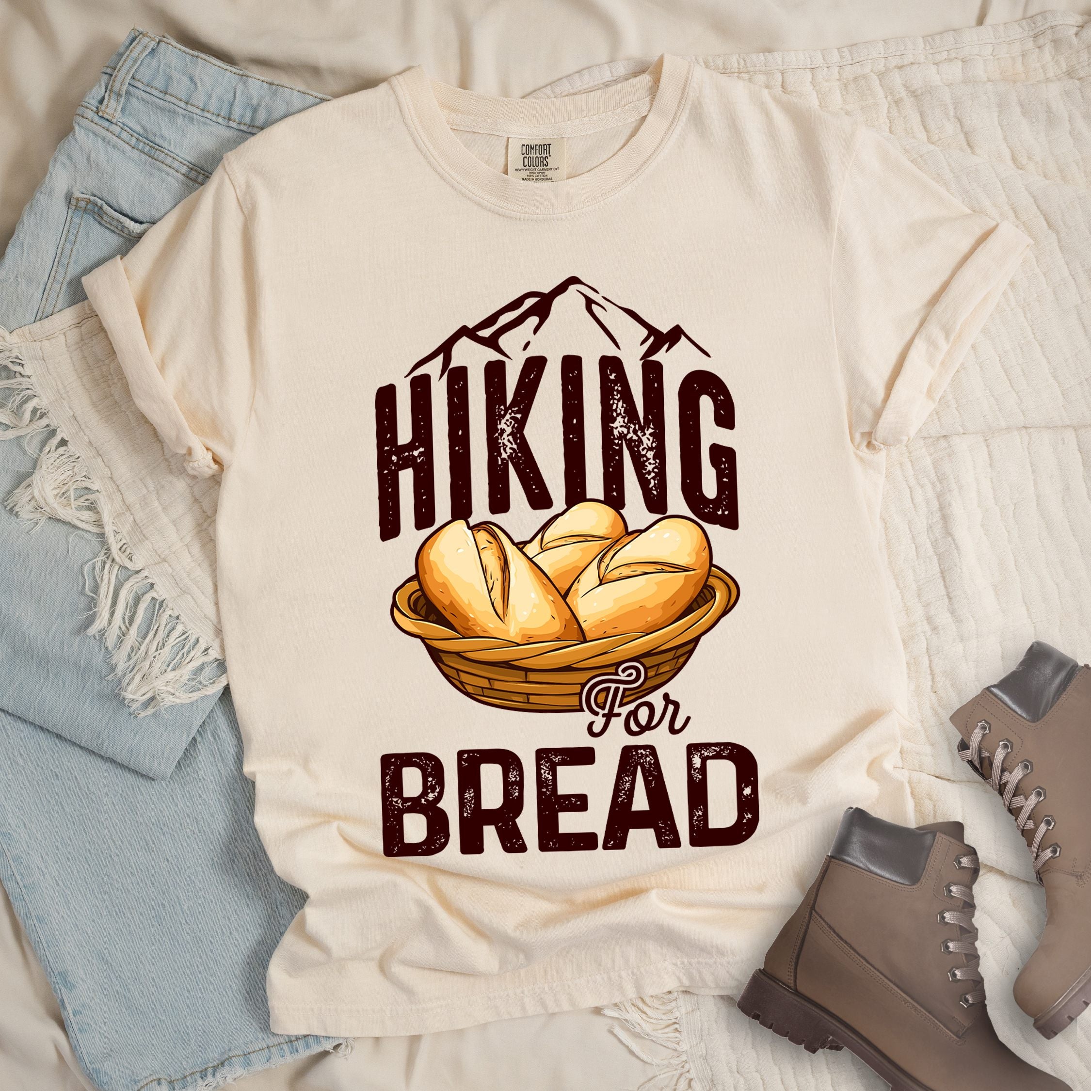 Ivory t-shirt with brown distressed text reading "HIKING For BREAD". "HIKING" appears in large brown distressed letters at the top with brown mountain outlines above it, "For" in small brown script in the middle, and "BREAD" in large brown distressed letters at the bottom. Features an illustration of bread rolls in a woven basket between the words "HIKING" and "BREAD". Features a classic crew neck and casual rolled sleeves, styled alongside light wash denim and brown hiking boots.