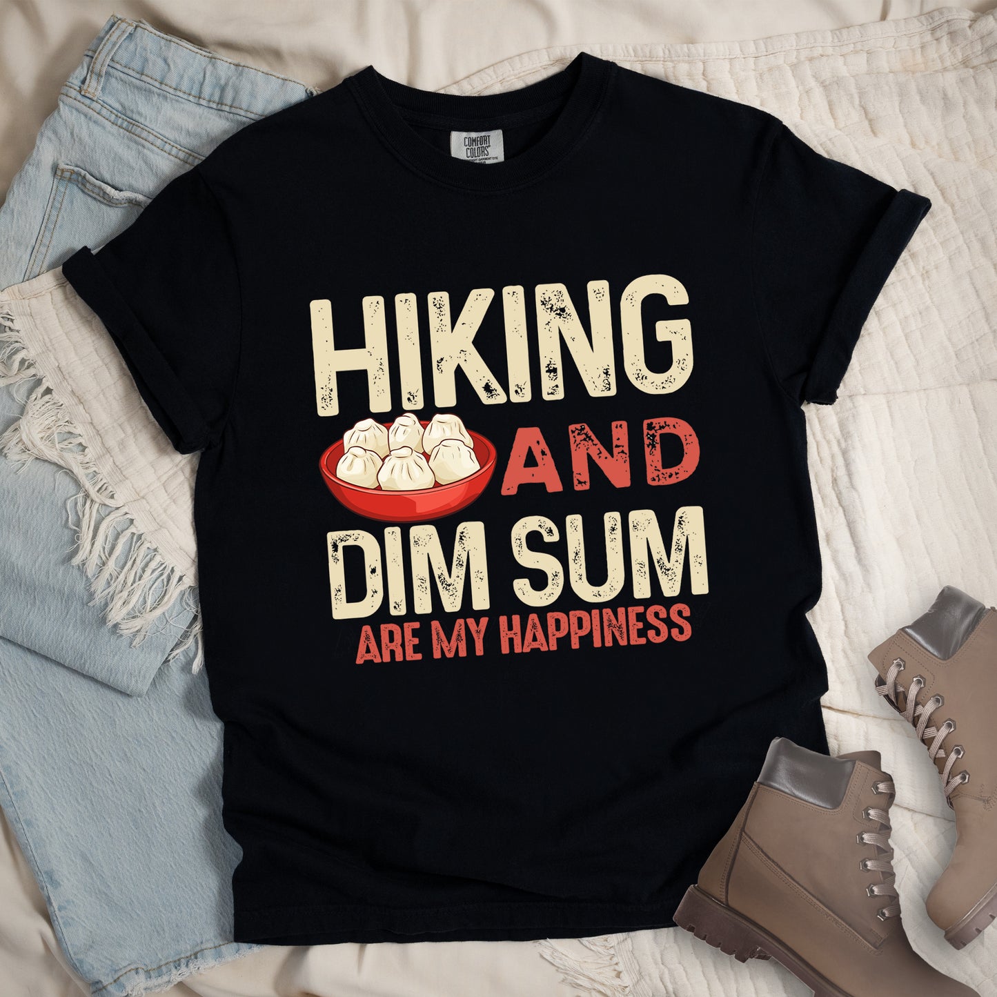 Black t-shirt text reading "HIKING AND DIM SUM ARE MY HAPPINESS". "HIKING" appears in large beige distressed letters at the top, "AND" in red distressed text in the middle, "DIM SUM" in large beige distressed letters below it, and "ARE MY HAPPINESS" in smaller red text at the bottom. Features an illustration of six steamed dumplings in a red bowl between "HIKING" and "AND". Features a classic crew neck and casual rolled sleeves, styled alongside light wash denim and brown hiking boots.