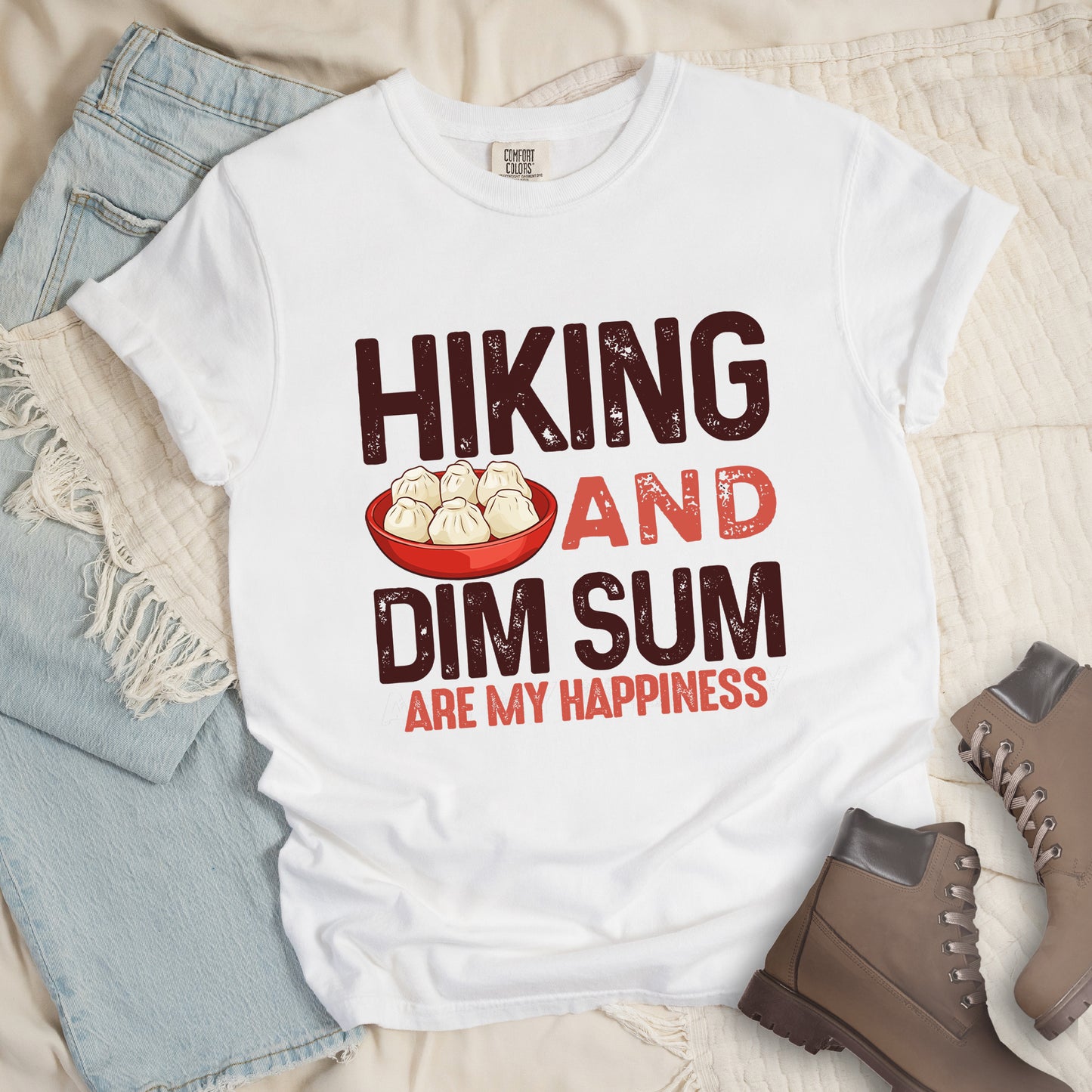White t-shirt with text reading "HIKING AND DIM SUM ARE MY HAPPINESS". "HIKING" appears in large brown distressed letters at the top, "AND" in red distressed text in the middle, "DIM SUM" in large brown distressed letters below it, and "ARE MY HAPPINESS" in smaller red text at the bottom. Features an illustration of six steamed dumplings in a red bowl between "HIKING" and "AND". Features a classic crew neck and casual rolled sleeves, styled alongside light wash denim and brown hiking boots.