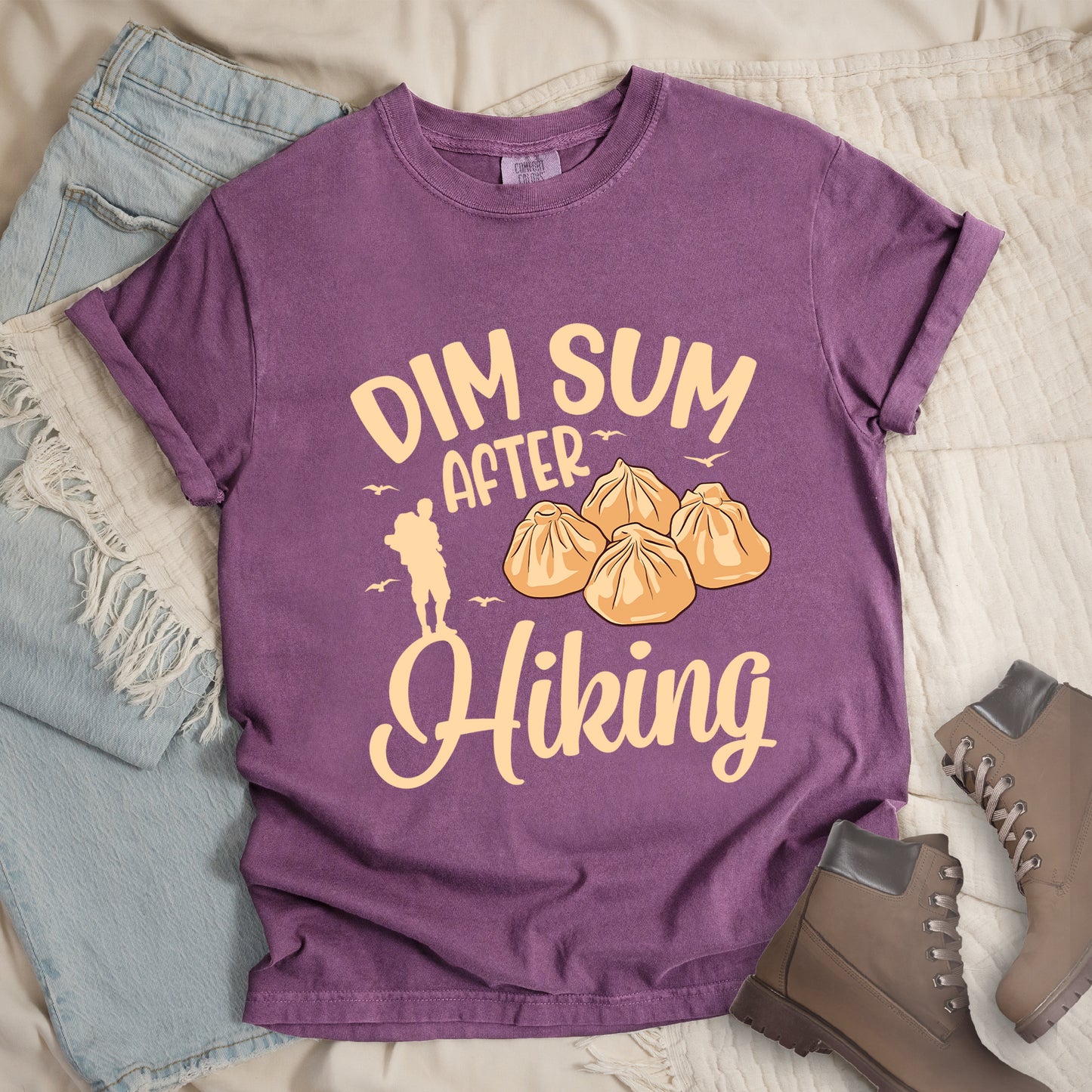 Berry (Dark Violet) t-shirt with beige text reading "DIM SUM AFTER Hiking". "DIM SUM" appears in large beige letters at the top, "AFTER" in smaller beige text in the middle, and "Hiking" in beige script at the bottom. Features an illustration of four steamed dumplings in the center, small beige flying birds, and a beige silhouette of a hiker to the left of "AFTER". Features a classic crew neck and casual rolled sleeves, styled alongside light wash denim and brown hiking boots.