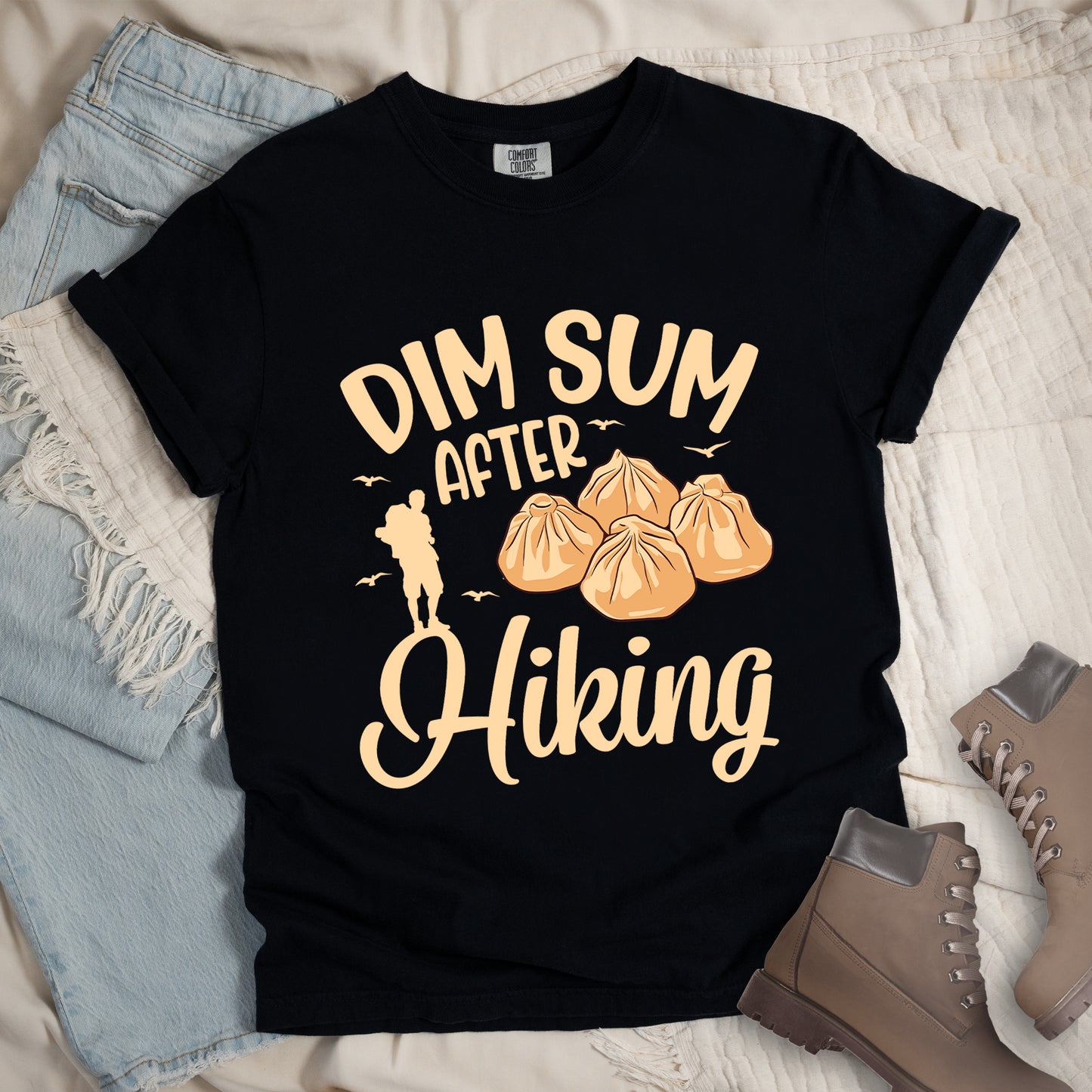 Black t-shirt with beige text reading "DIM SUM AFTER Hiking". "DIM SUM" appears in large beige letters at the top, "AFTER" in smaller beige text in the middle, and "Hiking" in beige script at the bottom. Features an illustration of four steamed dumplings in the center, small beige flying birds, and a beige silhouette of a hiker to the left of "AFTER". Features a classic crew neck and casual rolled sleeves, styled alongside light wash denim and brown hiking boots.