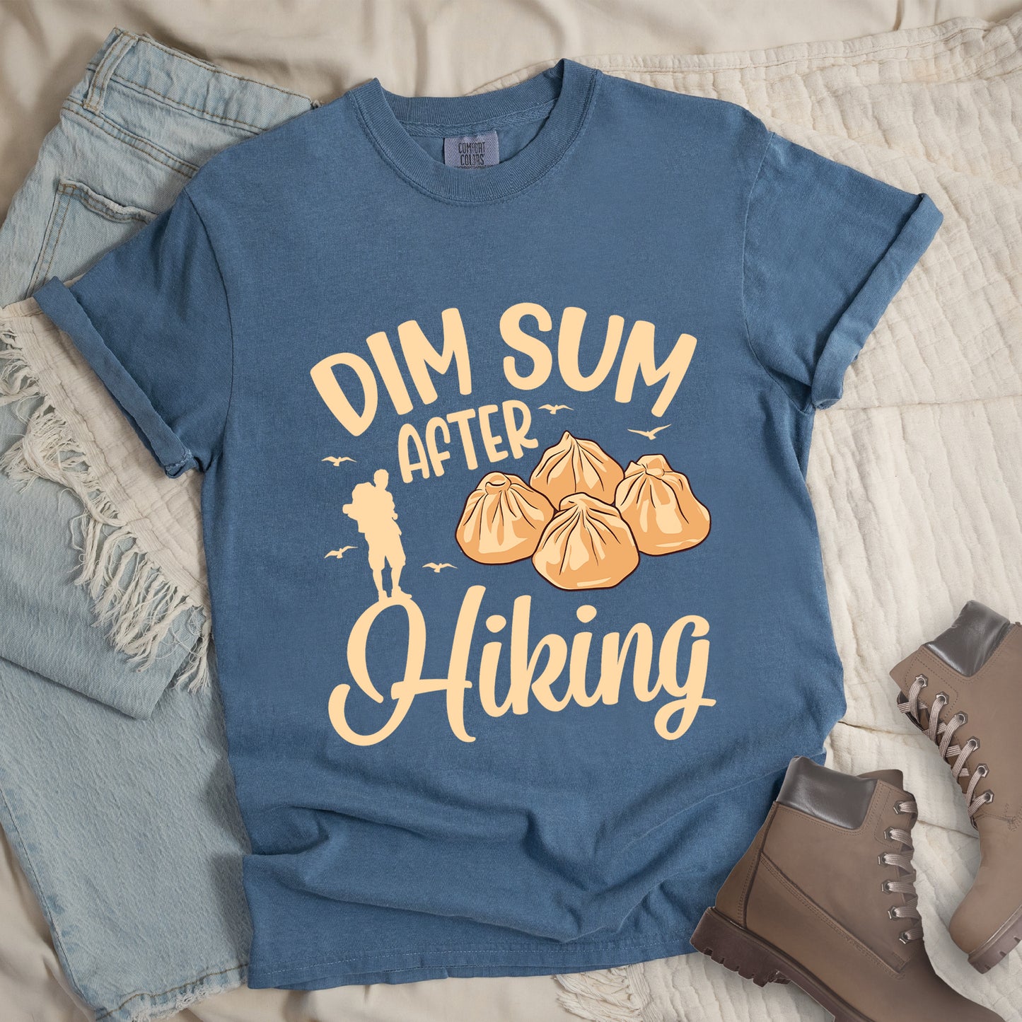 Blue Jean (Denim Blue) t-shirt with beige text reading "DIM SUM AFTER Hiking". "DIM SUM" appears in large beige letters at the top, "AFTER" in smaller beige text in the middle, and "Hiking" in beige script at the bottom. Features an illustration of four steamed dumplings in the center, small beige flying birds, and a beige silhouette of a hiker to the left of "AFTER". Features a classic crew neck and casual rolled sleeves, styled alongside light wash denim and brown hiking boots.