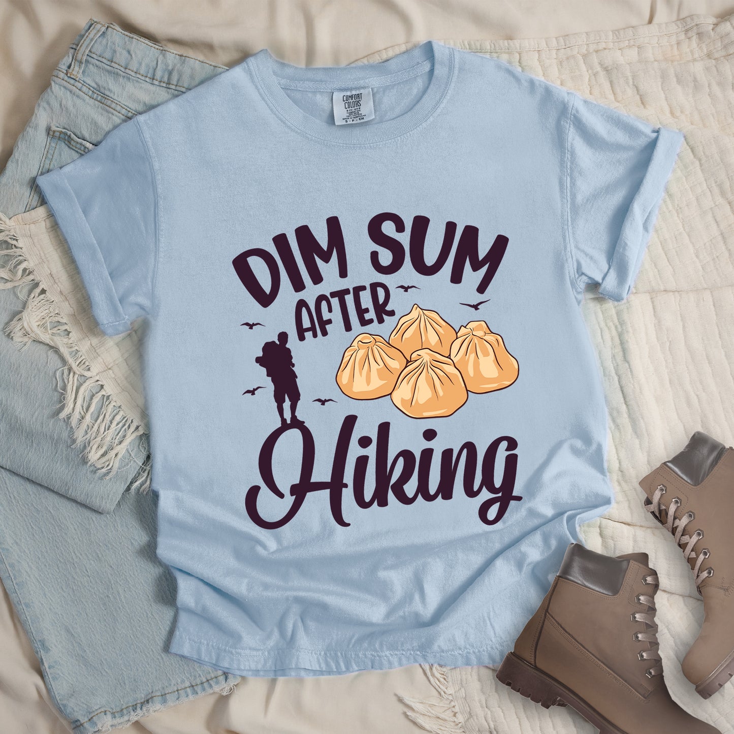Chambray (Light Blue) t-shirt with deep violet text reading "DIM SUM AFTER Hiking". "DIM SUM" appears in large deep violet letters at the top, "AFTER" in smaller deep violet text in the middle, and "Hiking" in deep violet script at the bottom. Features an illustration of four steamed dumplings in the center, small deep violet flying birds, and a deep violet silhouette of a hiker to the left of "AFTER". Features a classic crew neck and casual rolled sleeves, alongside light wash denim and brown hiking boots