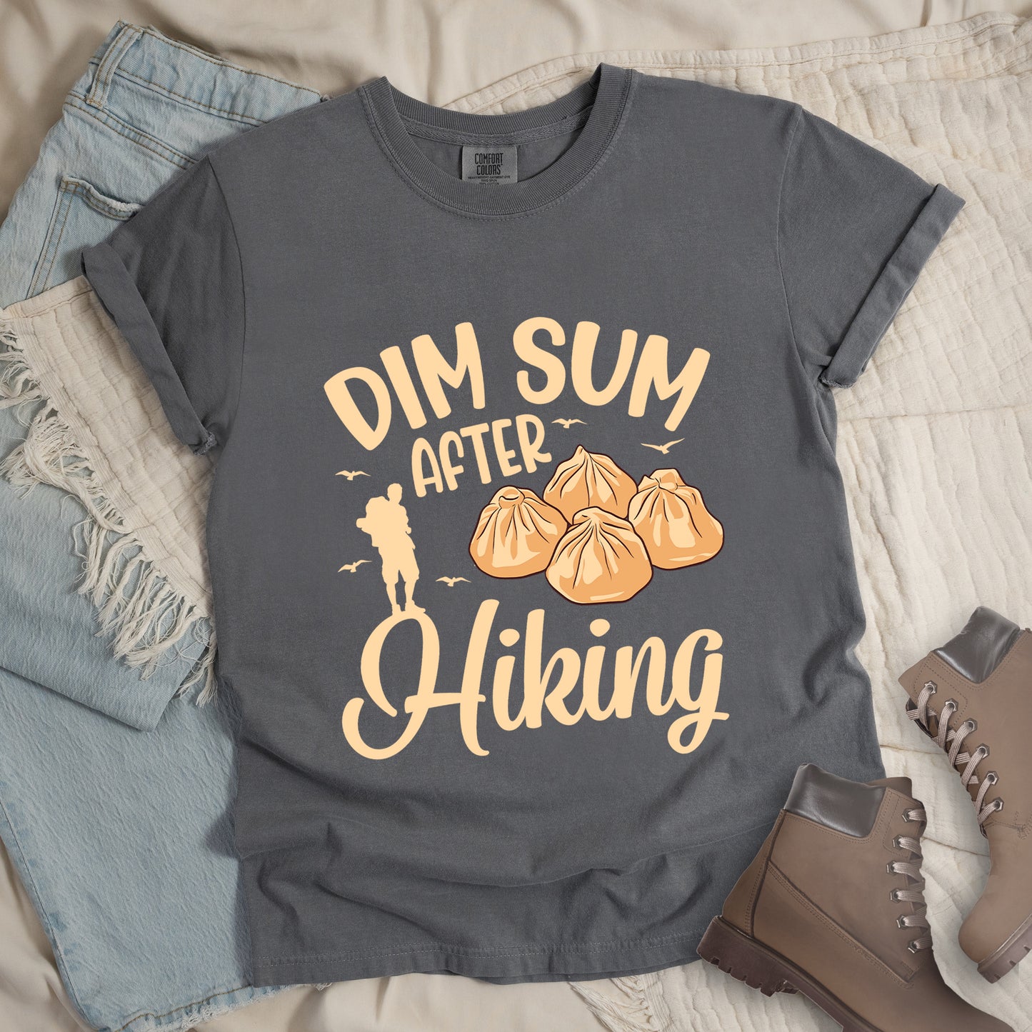 Grey t-shirt with beige text reading "DIM SUM AFTER Hiking". "DIM SUM" appears in large beige letters at the top, "AFTER" in smaller beige text in the middle, and "Hiking" in beige script at the bottom. Features an illustration of four steamed dumplings in the center, small beige flying birds, and a beige silhouette of a hiker to the left of "AFTER". Features a classic crew neck and casual rolled sleeves, styled alongside light wash denim and brown hiking boots.