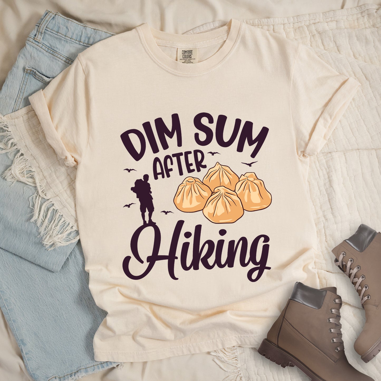 Ivory (Cream White) t-shirt with text reading "DIM SUM AFTER Hiking". "DIM SUM" appears in large black letters at the top, "AFTER" in smaller black text in the middle, and "Hiking" in black script at the bottom. Features an illustration of four steamed dumplings in the center, small deep violet flying birds, and a deep violet silhouette of a hiker to the left of "AFTER". Features a classic crew neck and casual rolled sleeves, styled alongside light wash denim and brown hiking boots.