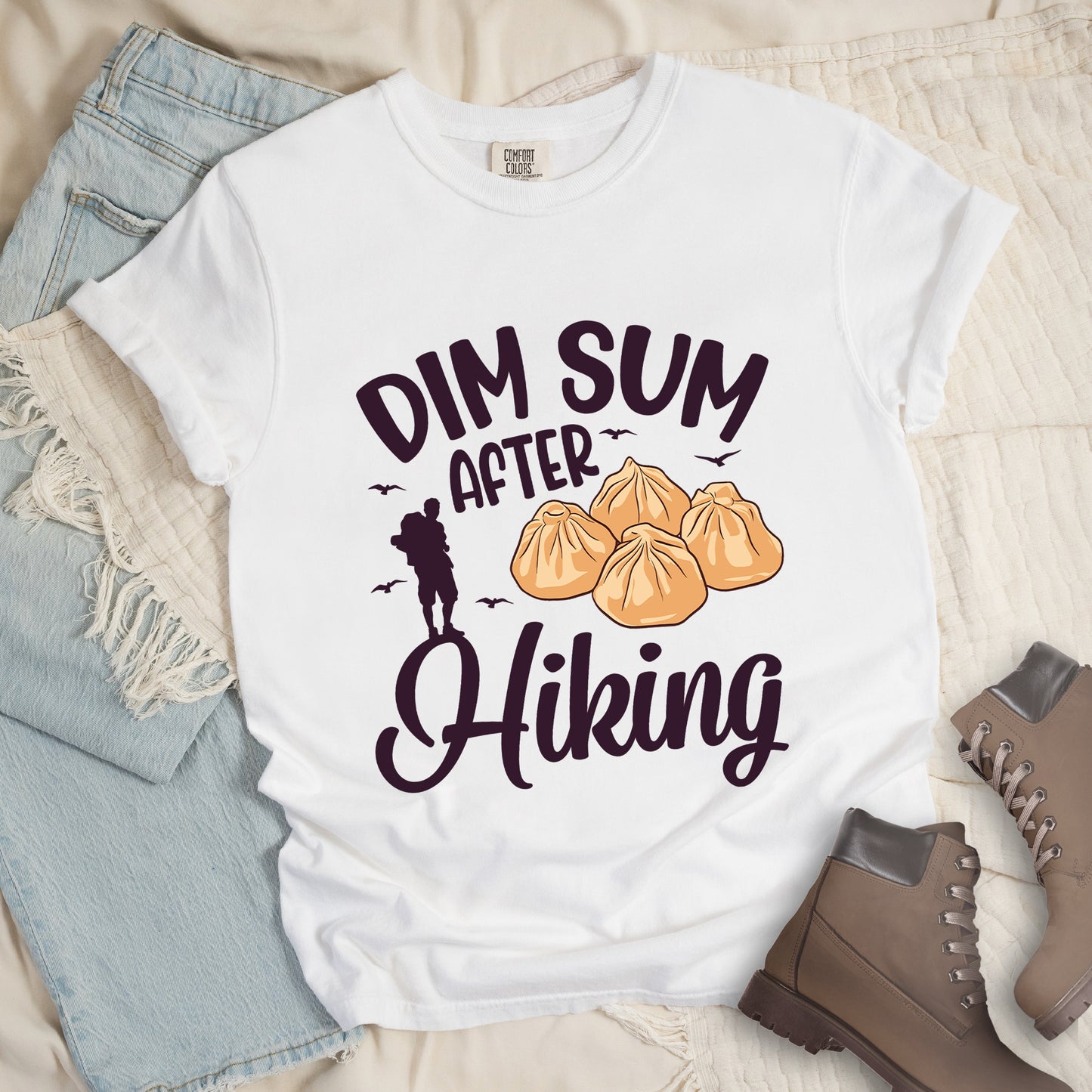 White t-shirt with deep violet text reading "DIM SUM AFTER Hiking". "DIM SUM" appears in large deep violet letters at the top, "AFTER" in smaller deep violet text in the middle, and "Hiking" in deep violet script at the bottom. Features an illustration of four steamed dumplings in the center, small deep violet flying birds, and a deep violet silhouette of a hiker to the left of "AFTER". Features a classic crew neck and casual rolled sleeves, styled alongside light wash denim and brown hiking boots.