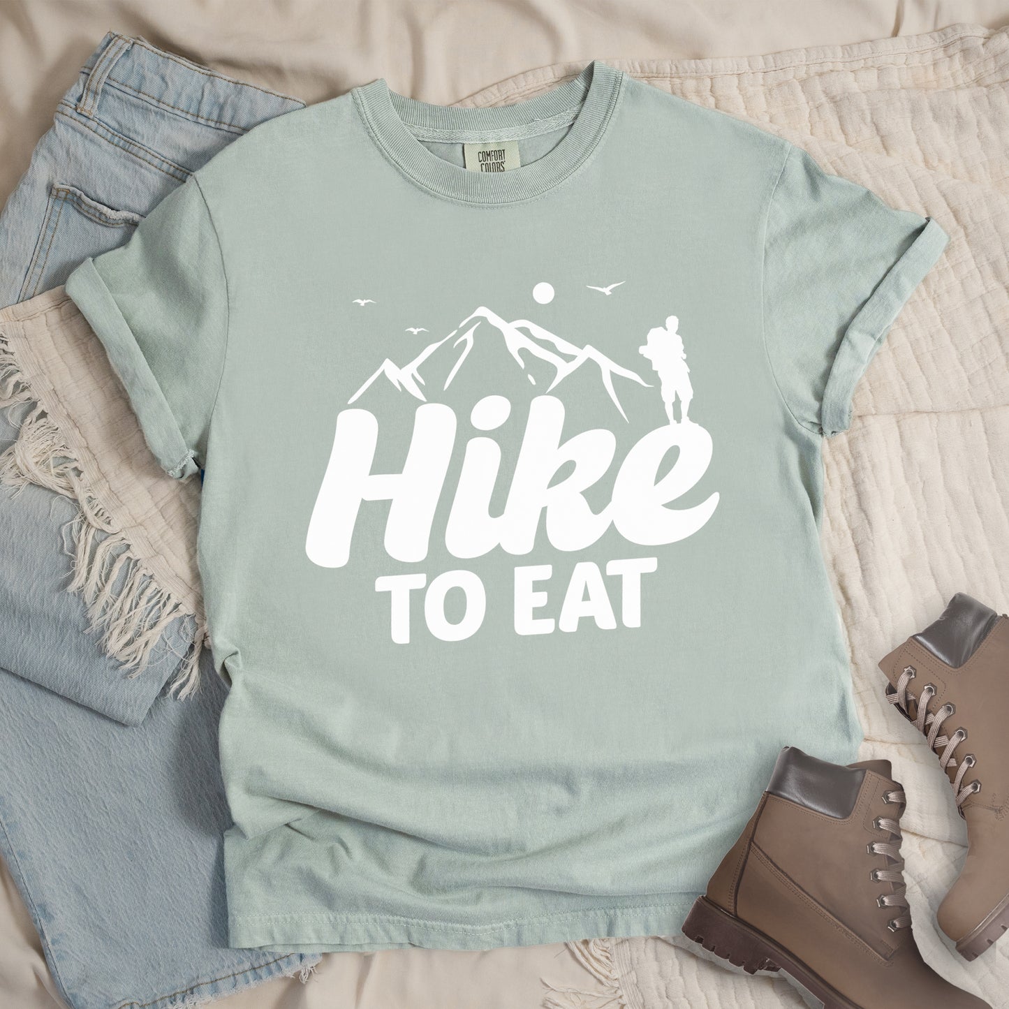 Bay (Light Gray) t-shirt with white text reading "Hike TO EAT". "Hike" appears in large white script letters with white mountain outlines above it, and "TO EAT" in bold white text below it. Features a white silhouette of a hiker to the right of the mountains, white flying birds, and a white circle representing the sun. Features a classic crew neck and casual rolled sleeves, styled alongside light wash denim and brown hiking boots.