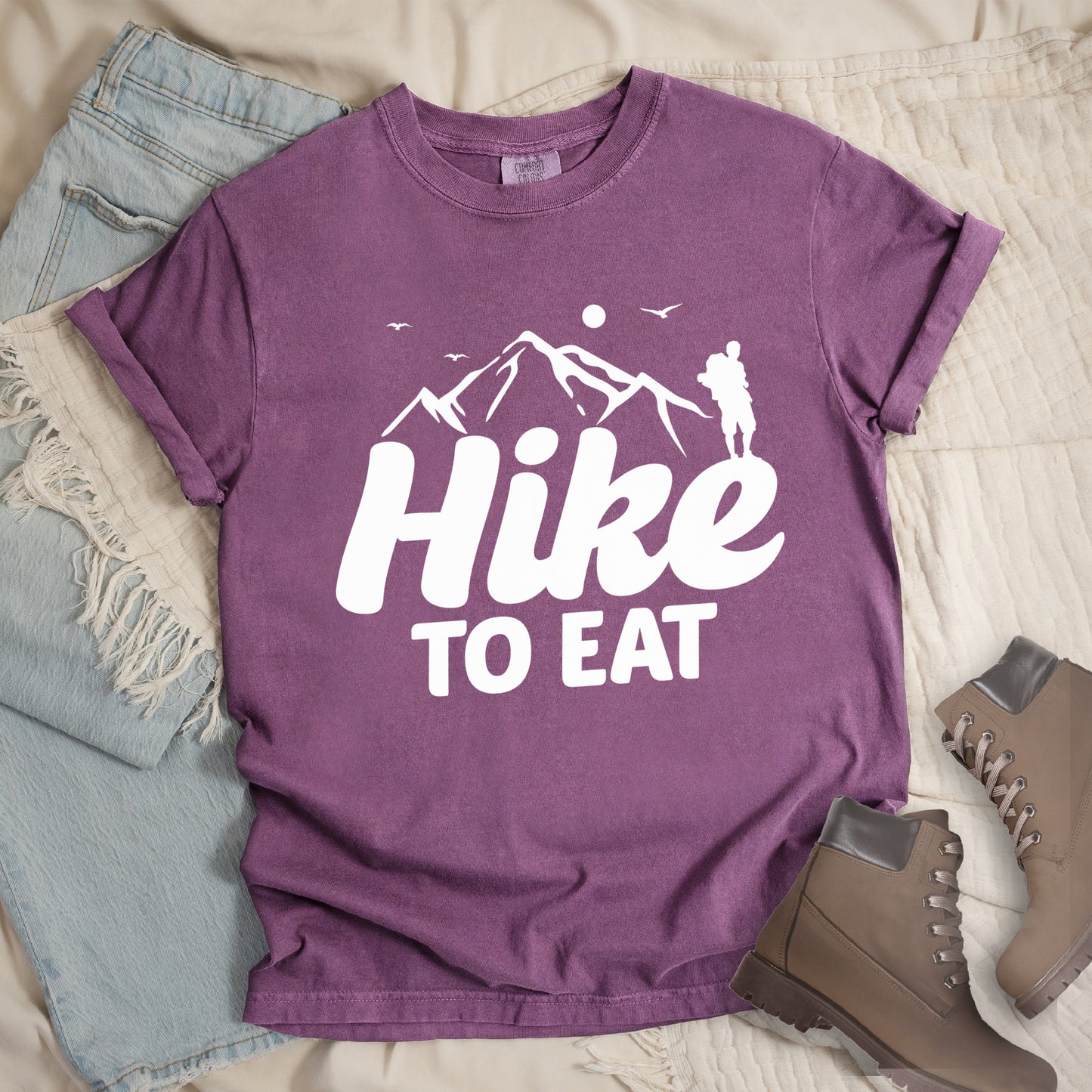 Berry (Dark Violet) t-shirt with white text reading "Hike TO EAT". "Hike" appears in large white script letters with white mountain outlines above it, and "TO EAT" in bold white text below it. Features a white silhouette of a hiker to the right of the mountains, white flying birds, and a white circle representing the sun. Features a classic crew neck and casual rolled sleeves, styled alongside light wash denim and brown hiking boots.