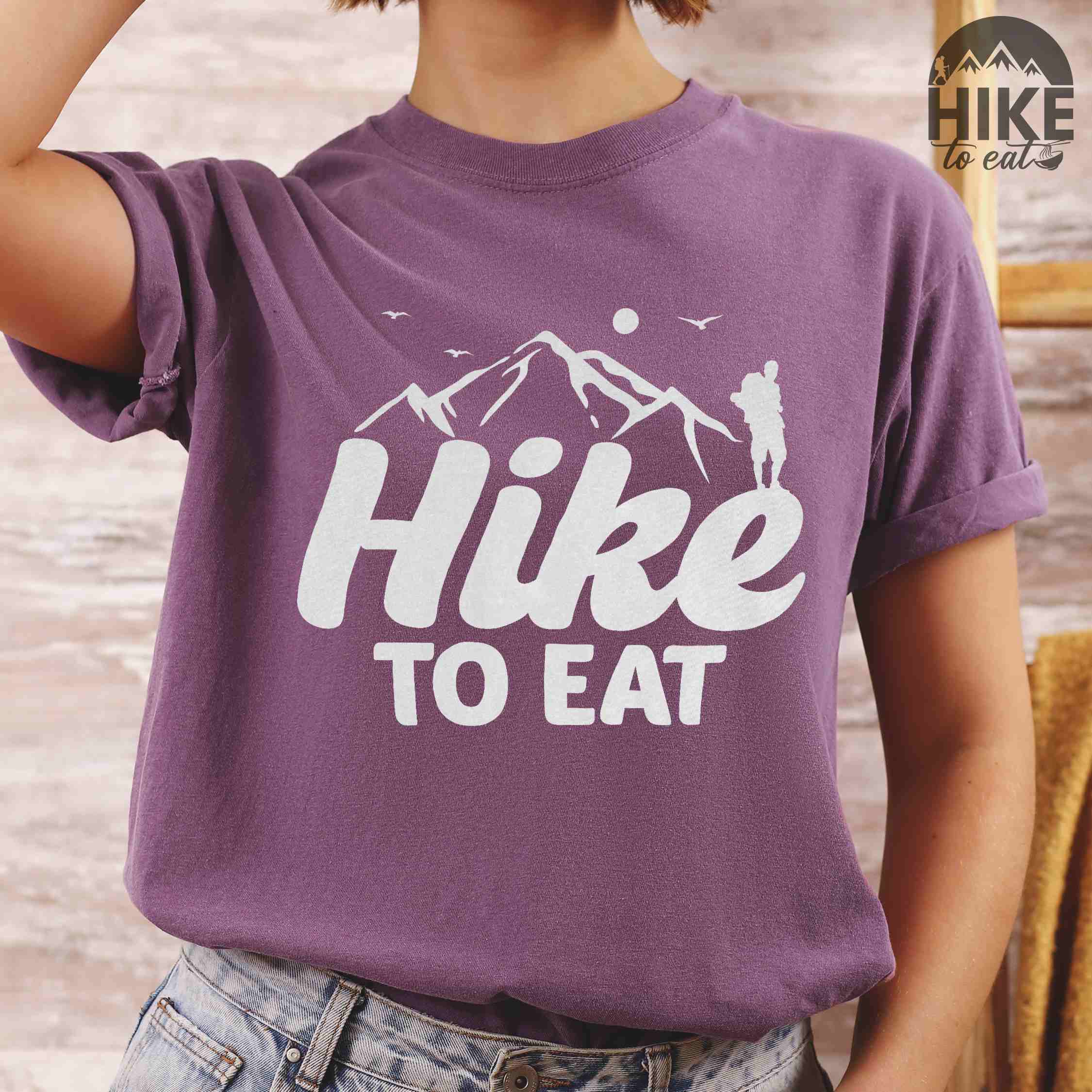 woman wearing berry "Hike to Eat" colored statement tee with bold typography, mountains, birds, sunrise, perfect for food motivated individuals.