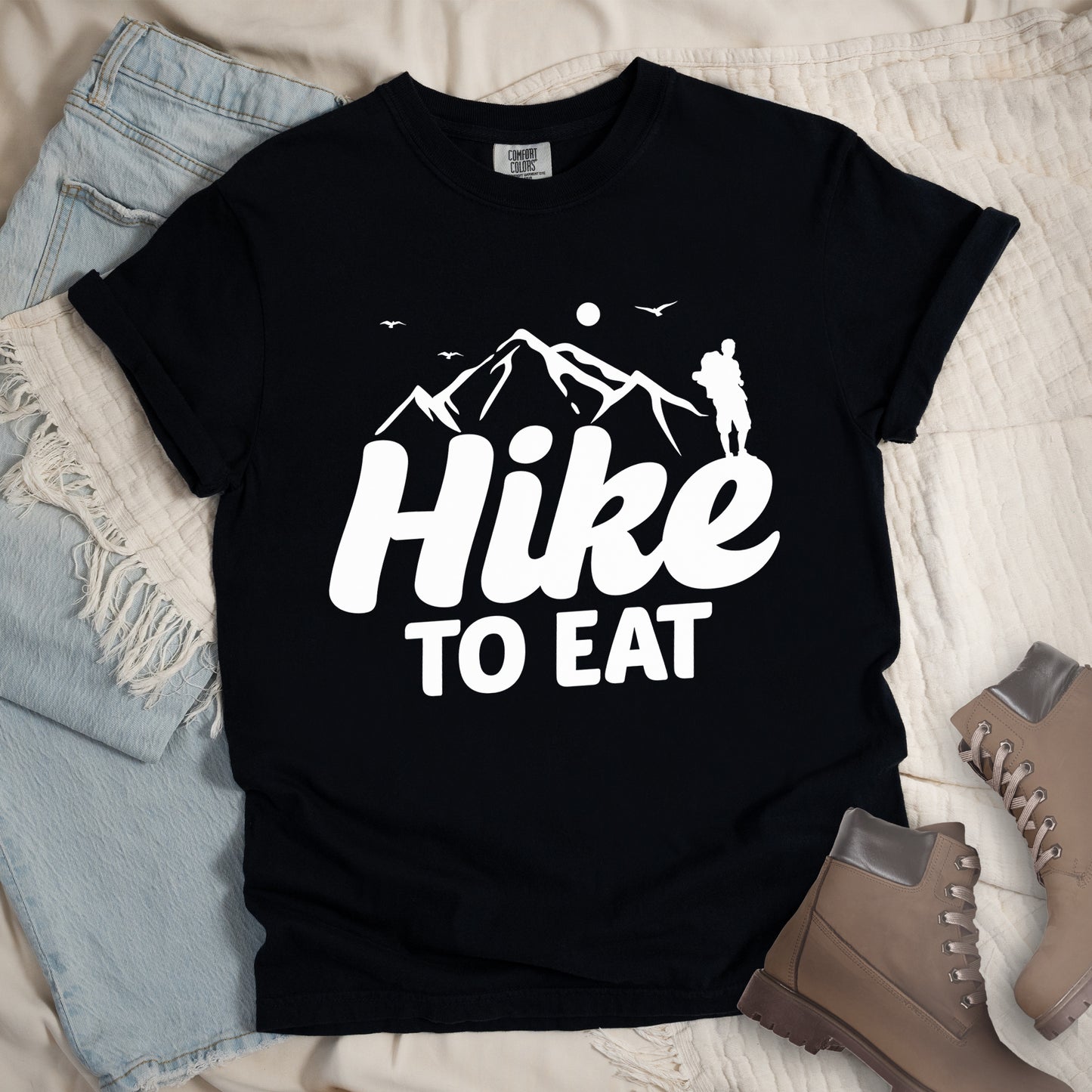 Black t-shirt with white text reading "Hike TO EAT". "Hike" appears in large white script letters with white mountain outlines above it, and "TO EAT" in bold white text below it. Features a white silhouette of a hiker to the right of the mountains, white flying birds, and a white circle representing the sun. Features a classic crew neck and casual rolled sleeves, styled alongside light wash denim and brown hiking boots.