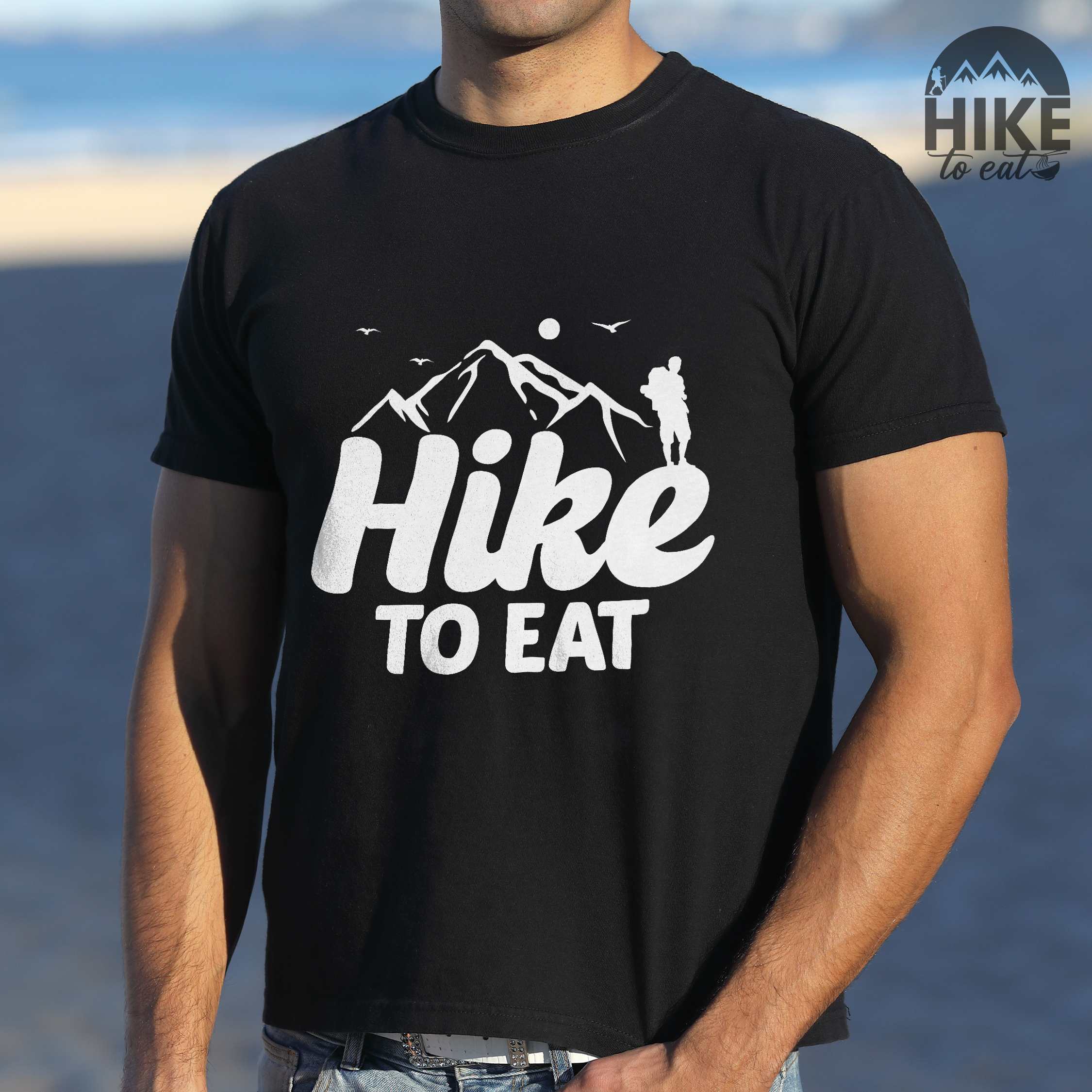 Man wearing black "Hike to Eat" colored statement tee with bold typography, mountains, birds, sunrise, perfect for food motivated individuals.