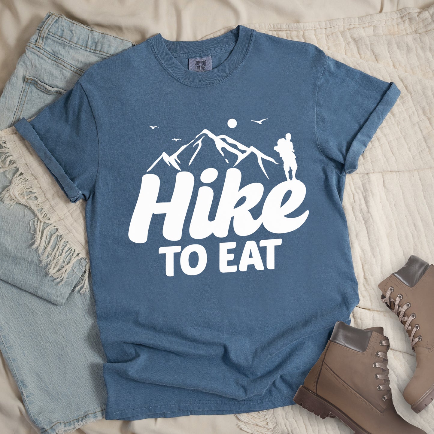 Blue Jean (Denim Blue) t-shirt with white text reading "Hike TO EAT". "Hike" appears in large white script letters with white mountain outlines above it, and "TO EAT" in bold white text below it. Features a white silhouette of a hiker to the right of the mountains, white flying birds, and a white circle representing the sun. Features a classic crew neck and casual rolled sleeves, styled alongside light wash denim and brown hiking boots.