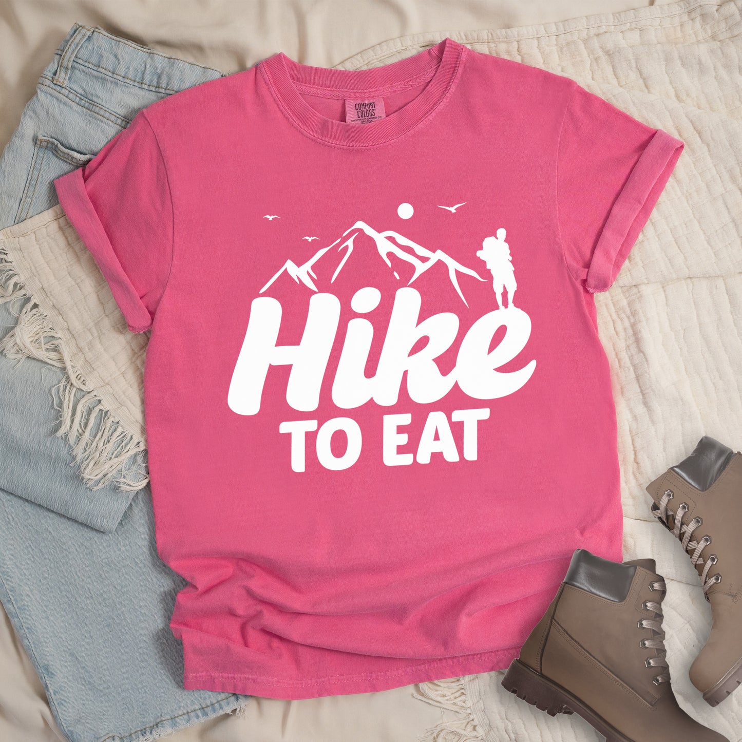 Crunchberry (Bright Pink) t-shirt with white text reading "Hike TO EAT". "Hike" appears in large white script letters with white mountain outlines above it, and "TO EAT" in bold white text below it. Features a white silhouette of a hiker to the right of the mountains, white flying birds, and a white circle representing the sun. Features a classic crew neck and casual rolled sleeves, styled alongside light wash denim and brown hiking boots.