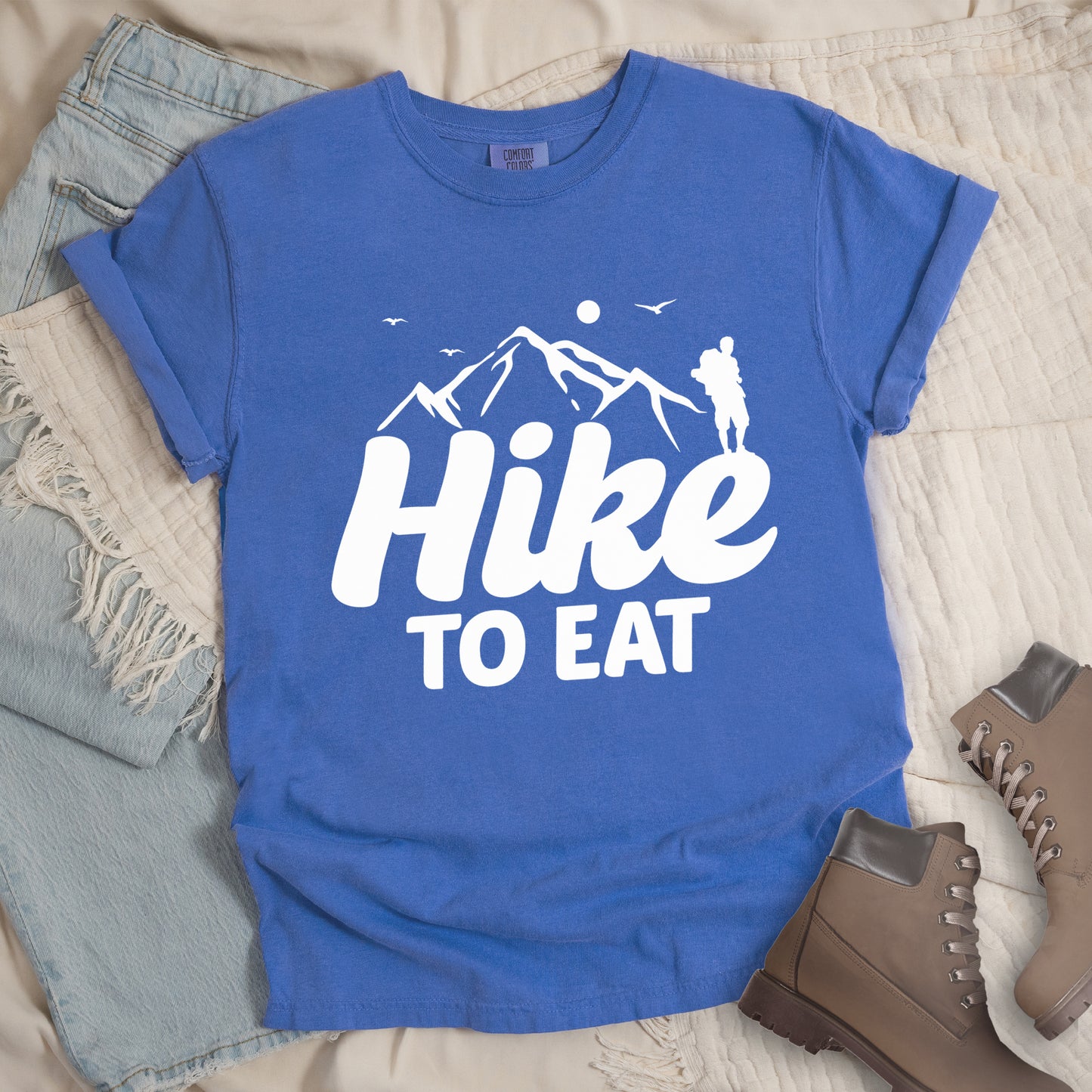 Flo Blue (Bright Blue) t-shirt with white text reading "Hike TO EAT". "Hike" appears in large white script letters with white mountain outlines above it, and "TO EAT" in bold white text below it. Features a white silhouette of a hiker to the right of the mountains, white flying birds, and a white circle representing the sun. Features a classic crew neck and casual rolled sleeves, styled alongside light wash denim and brown hiking boots.