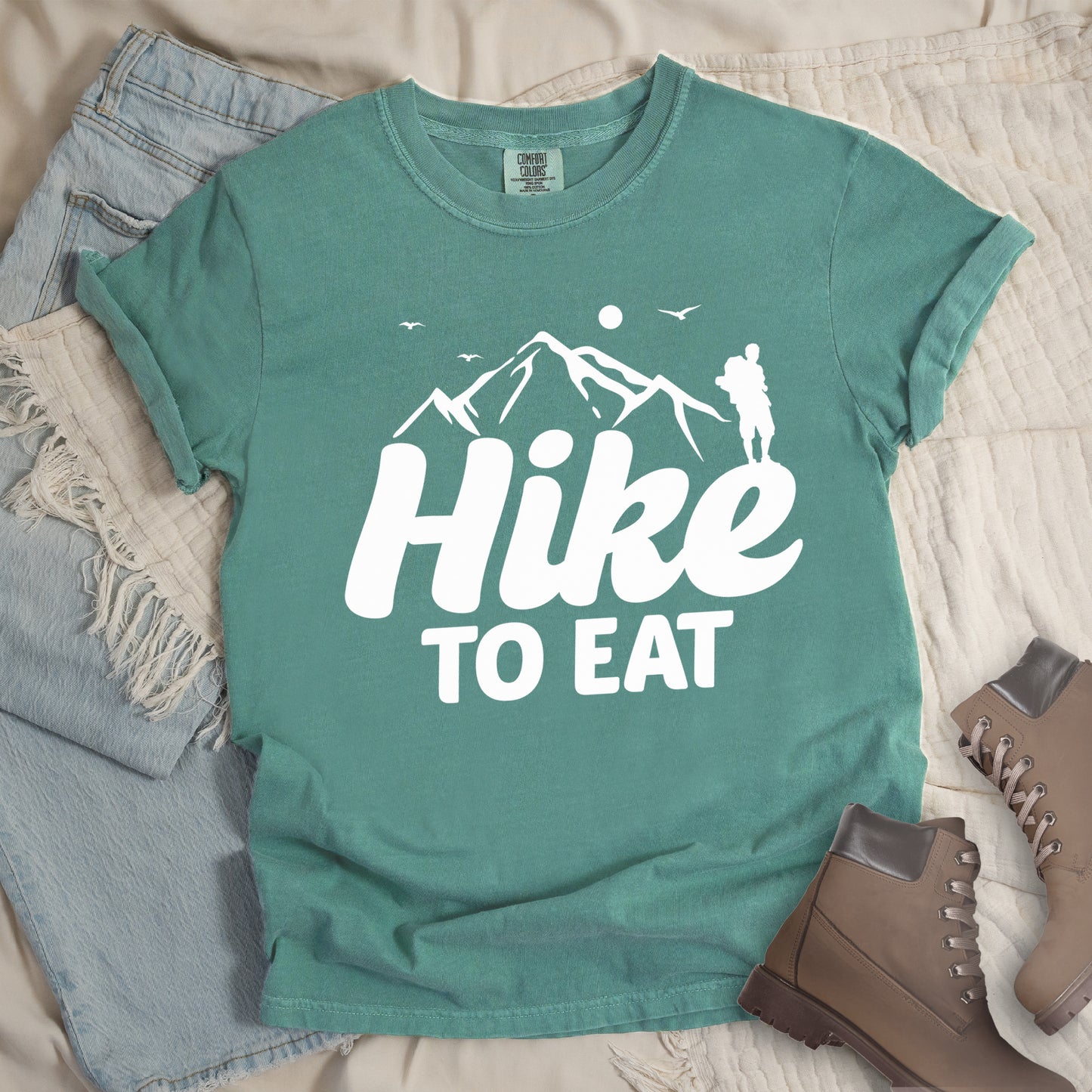 Light Green t-shirt with white text reading "Hike TO EAT". "Hike" appears in large white script letters with white mountain outlines above it, and "TO EAT" in bold white text below it. Features a white silhouette of a hiker to the right of the mountains, white flying birds, and a white circle representing the sun. Features a classic crew neck and casual rolled sleeves, styled alongside light wash denim and brown hiking boots.