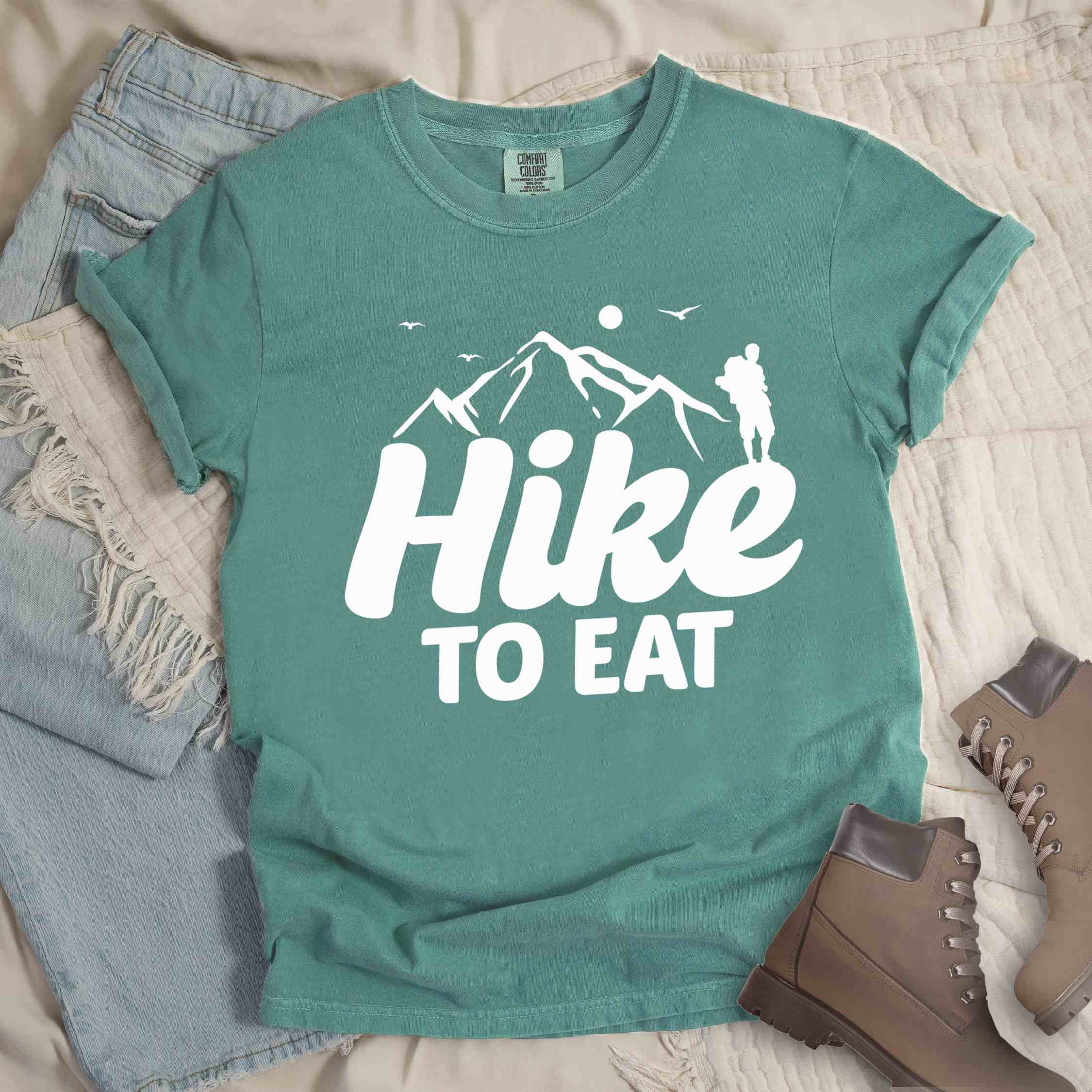 light green "Hike to Eat" colored statement tee with bold typography, mountains, birds, sunrise, perfect for food motivated individuals.