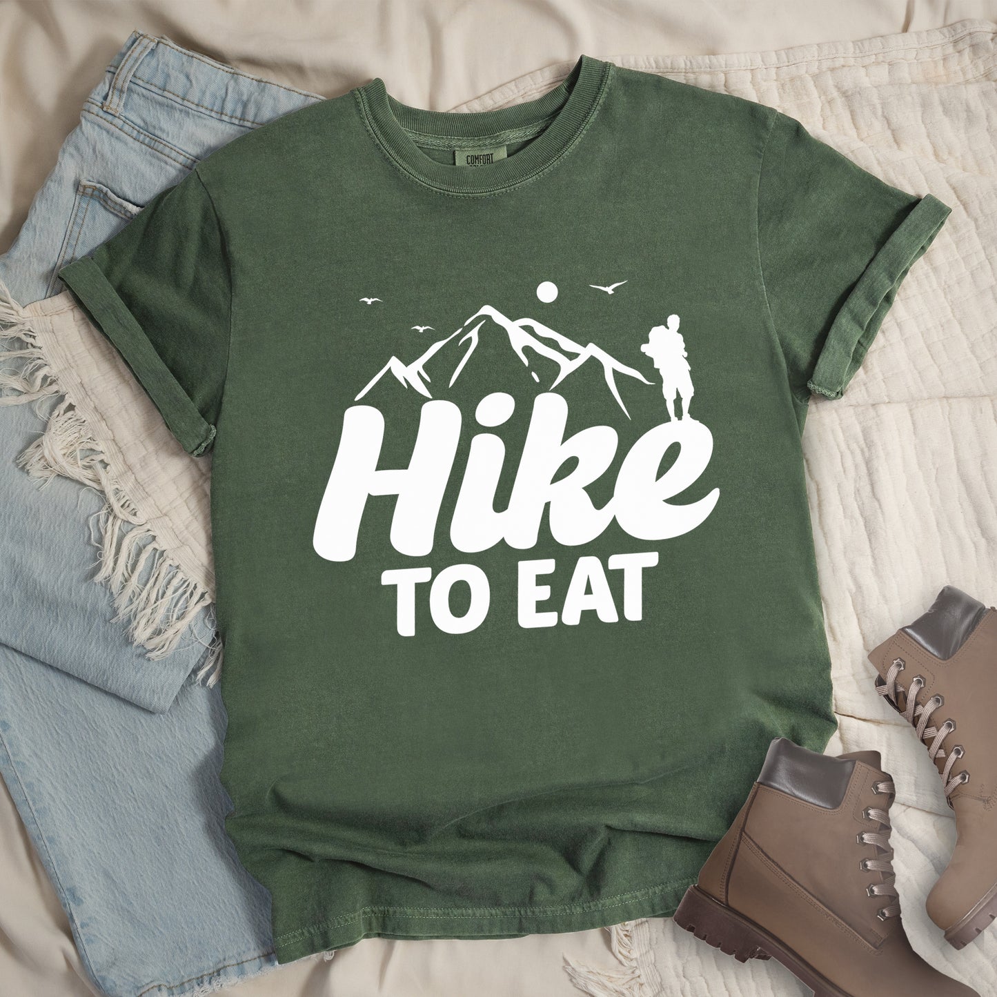 Moss (Dark Green) t-shirt with white text reading "Hike TO EAT". "Hike" appears in large white script letters with white mountain outlines above it, and "TO EAT" in bold white text below it. Features a white silhouette of a hiker to the right of the mountains, white flying birds, and a white circle representing the sun. Features a classic crew neck and casual rolled sleeves, styled alongside light wash denim and brown hiking boots.