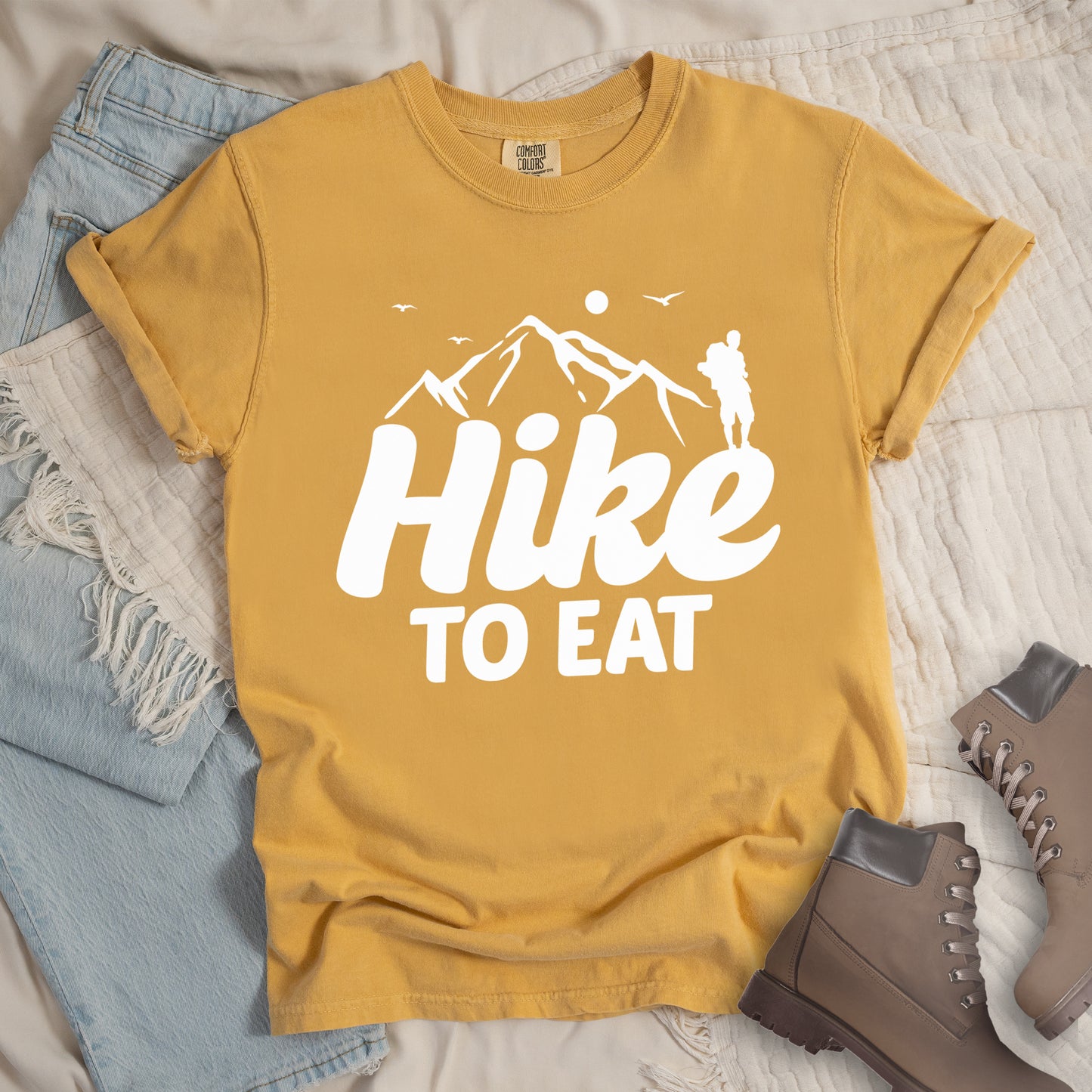 Mustard (Yellow) t-shirt with white text reading "Hike TO EAT". "Hike" appears in large white script letters with white mountain outlines above it, and "TO EAT" in bold white text below it. Features a white silhouette of a hiker to the right of the mountains, white flying birds, and a white circle representing the sun. Features a classic crew neck and casual rolled sleeves, styled alongside light wash denim and brown hiking boots.