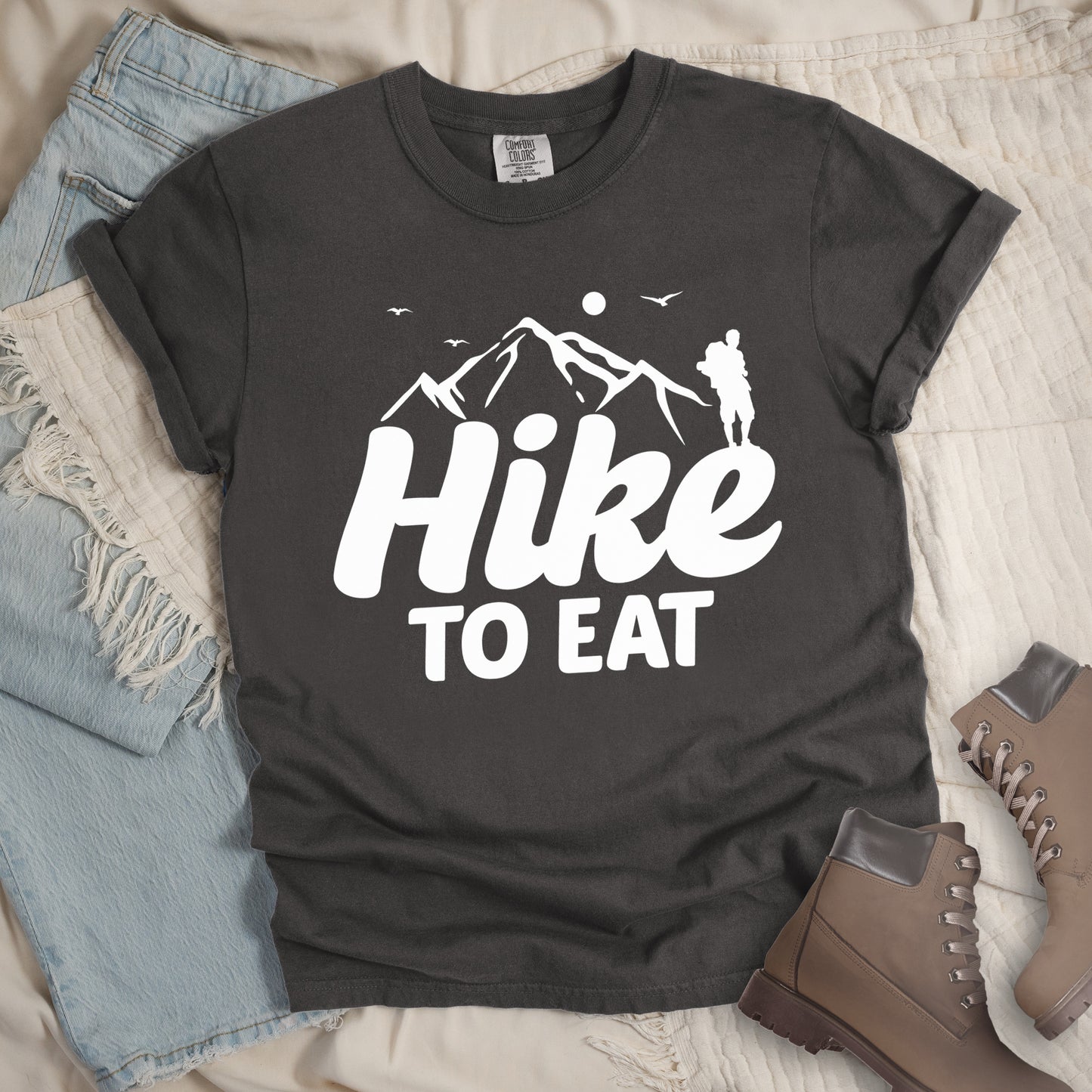 Pepper (Dark Gray) t-shirt with white text reading "Hike TO EAT". "Hike" appears in large white script letters with white mountain outlines above it, and "TO EAT" in bold white text below it. Features a white silhouette of a hiker to the right of the mountains, white flying birds, and a white circle representing the sun. Features a classic crew neck and casual rolled sleeves, styled alongside light wash denim and brown hiking boots.