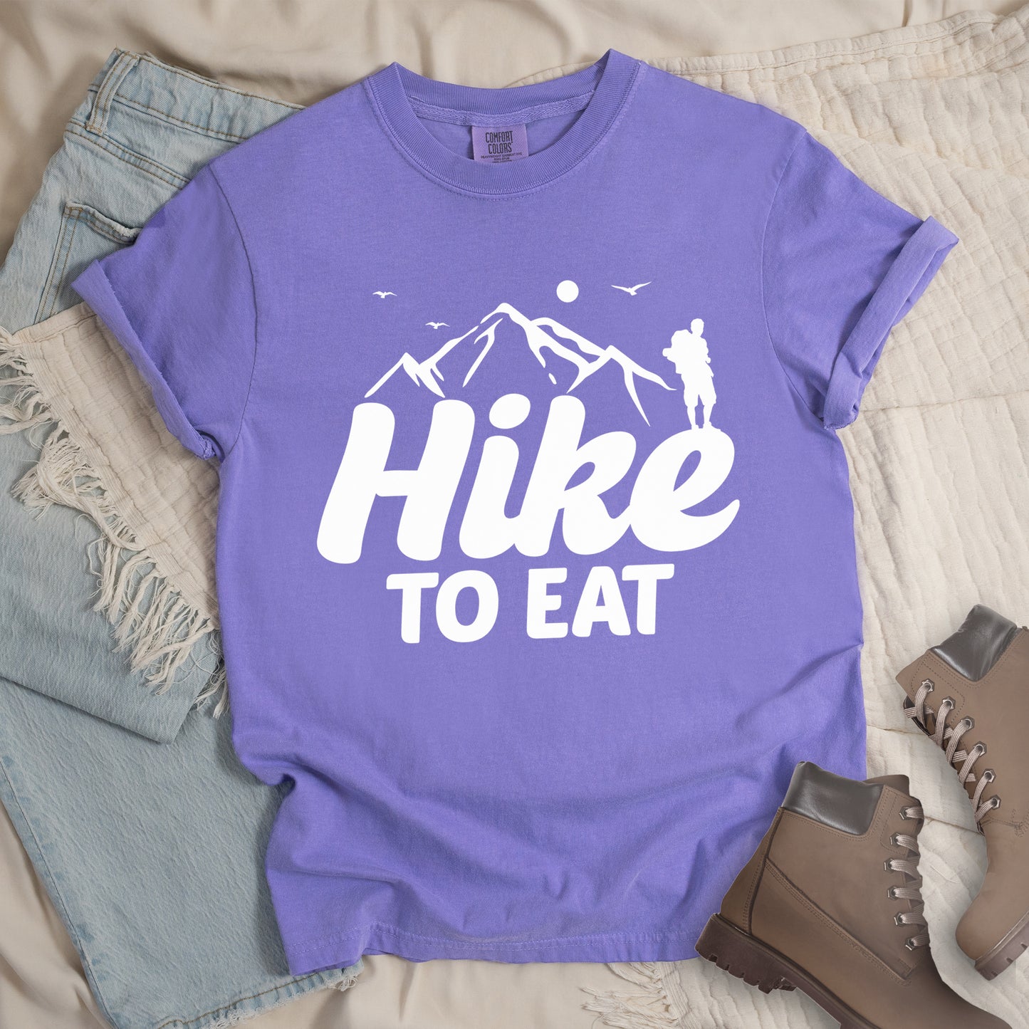 Violet t-shirt with white text reading "Hike TO EAT". "Hike" appears in large white script letters with white mountain outlines above it, and "TO EAT" in bold white text below it. Features a white silhouette of a hiker to the right of the mountains, white flying birds, and a white circle representing the sun. Features a classic crew neck and casual rolled sleeves, styled alongside light wash denim and brown hiking boots.
