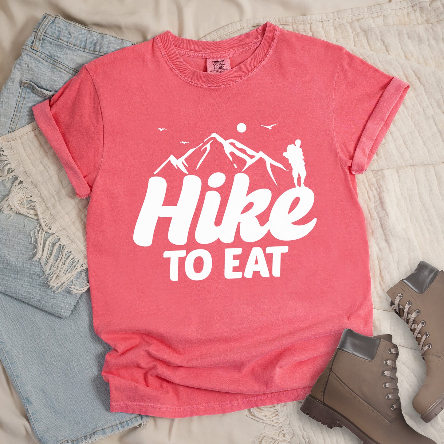 Watermelon (Light Coral) t-shirt with white text reading "Hike TO EAT". "Hike" appears in large white script letters with white mountain outlines above it, and "TO EAT" in bold white text below it. Features a white silhouette of a hiker to the right of the mountains, white flying birds, and a white circle representing the sun. Features a classic crew neck and casual rolled sleeves, styled alongside light wash denim and brown hiking boots.