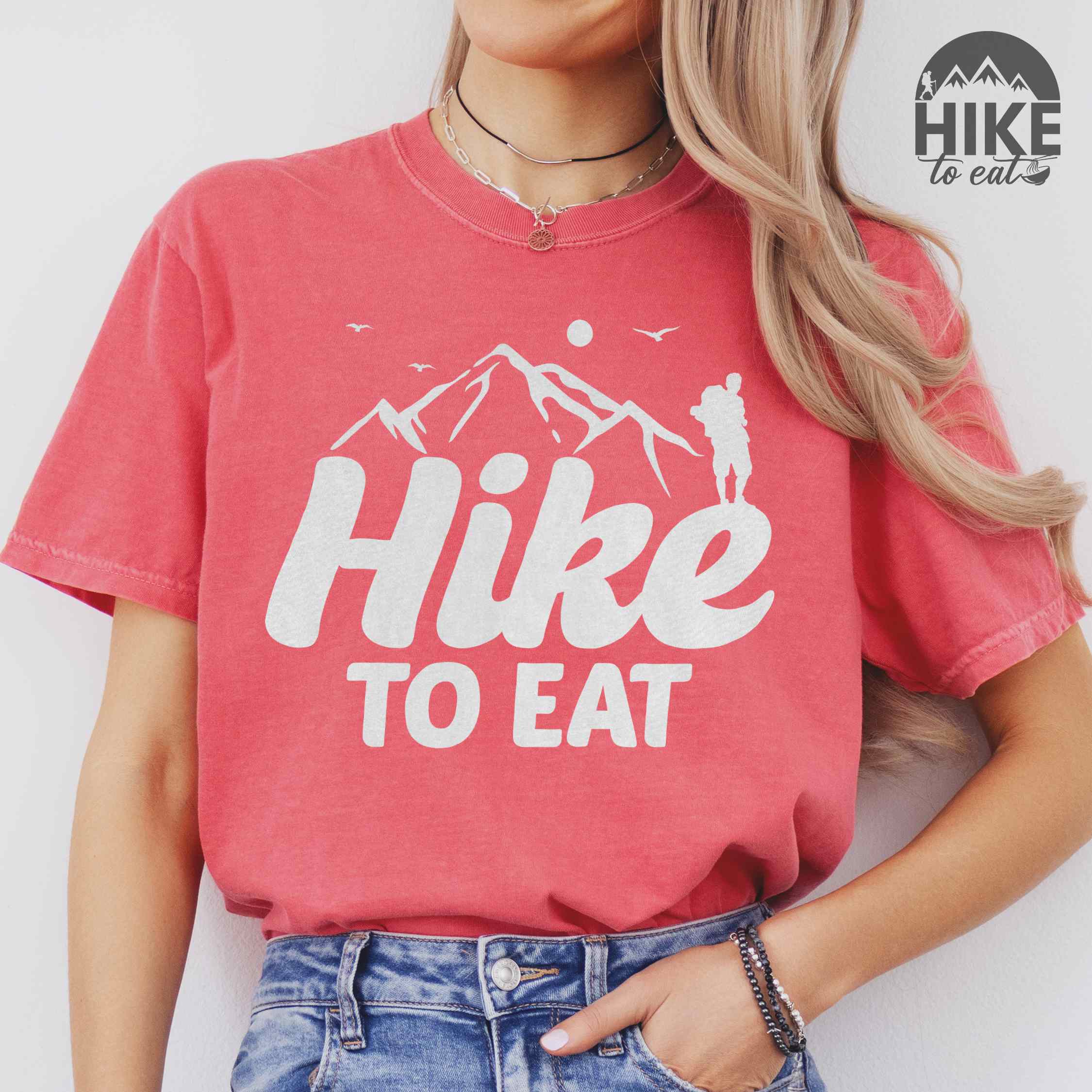 woman wearing watermelon "Hike to Eat" colored statement tee with bold typography, mountains, birds, sunrise, perfect for food motivated individuals.