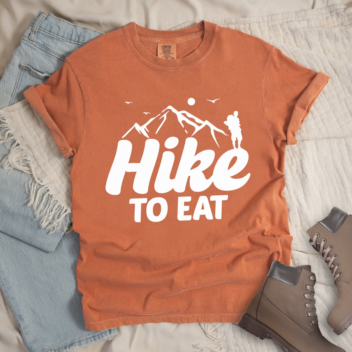 Yam (Brown) t-shirt with white text reading "Hike TO EAT". "Hike" appears in large white script letters with white mountain outlines above it, and "TO EAT" in bold white text below it. Features a white silhouette of a hiker to the right of the mountains, white flying birds, and a white circle representing the sun. Features a classic crew neck and casual rolled sleeves, styled alongside light wash denim and brown hiking boots.