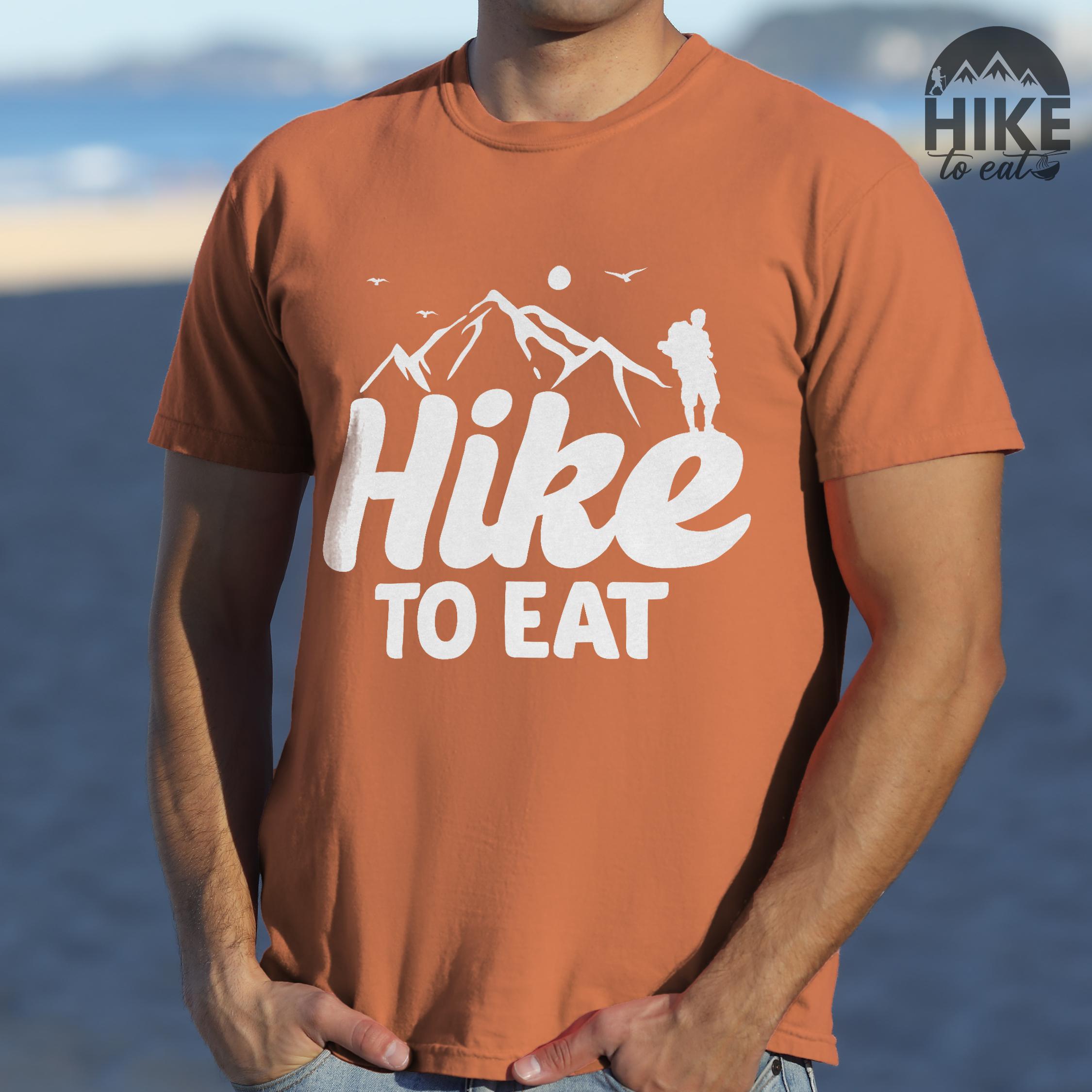 man wearing yam "Hike to Eat" colored statement tee with bold typography, mountains, birds, sunrise, perfect for food motivated individuals.