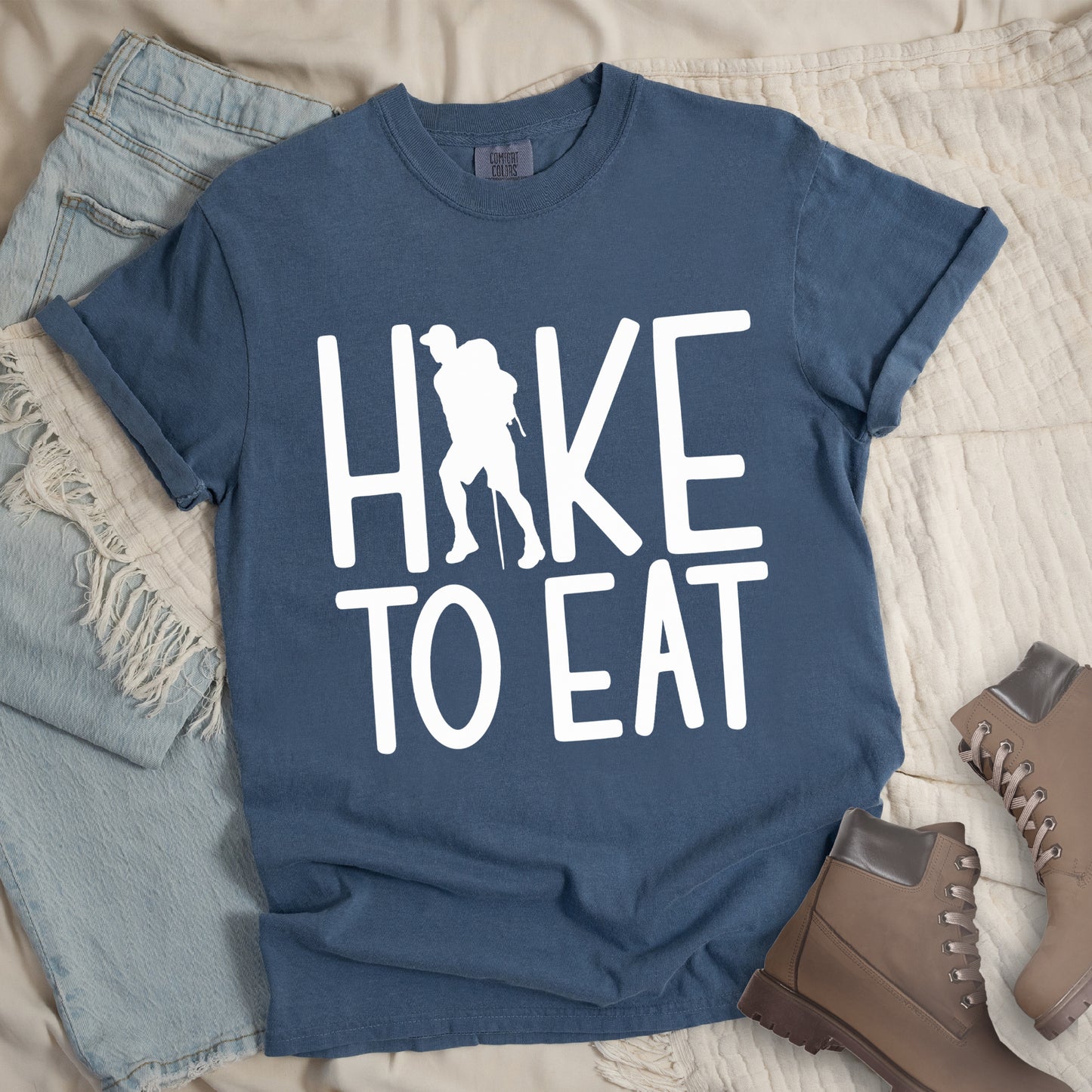 Blue Jean (Denim Blue) t-shirt with white text reading "HIKE TO EAT". "HIKE" appears in large white letters at the top with a white silhouette of a hiker with a backpack and walking stick integrated into the letter "I", and "TO EAT" in white text below it. The shirt features a classic crew neck and casual rolled sleeves, styled alongside light wash denim and brown hiking boots.
