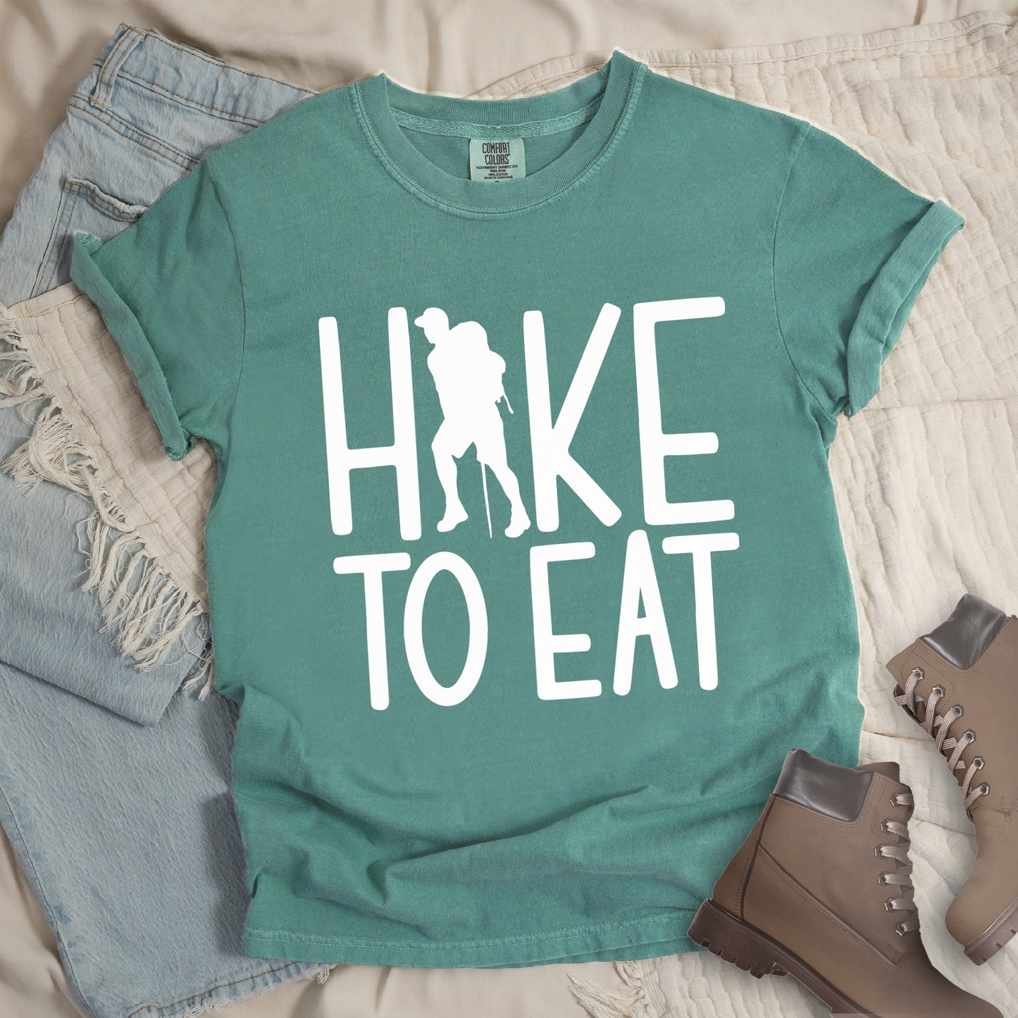 Light Green t-shirt with white text reading "HIKE TO EAT". "HIKE" appears in large white letters at the top with a white silhouette of a hiker with a backpack and walking stick integrated into the letter "I", and "TO EAT" in white text below it. The shirt features a classic crew neck and casual rolled sleeves, styled alongside light wash denim and brown hiking boots.