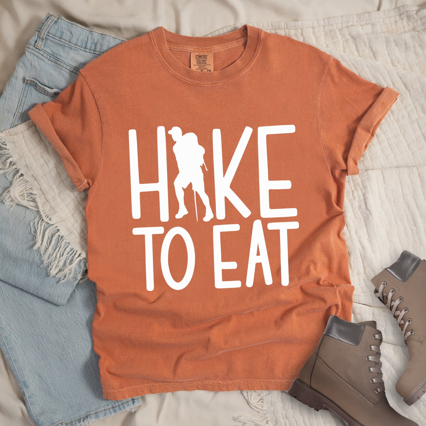 Yam (Brown) t-shirt with white text reading "HIKE TO EAT". "HIKE" appears in large white letters at the top with a white silhouette of a hiker with a backpack and walking stick integrated into the letter "I", and "TO EAT" in white text below it. The shirt features a classic crew neck and casual rolled sleeves, styled alongside light wash denim and brown hiking boots.