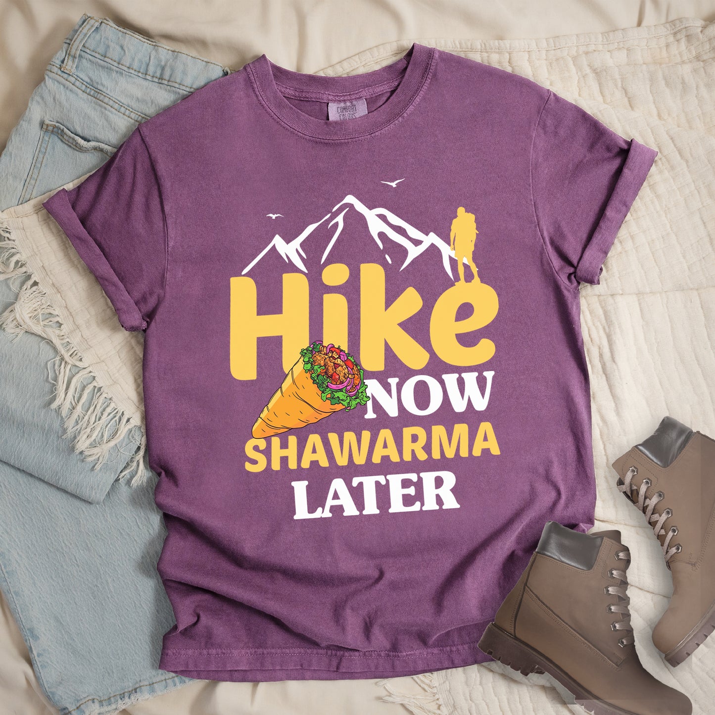 Berry (Dark Violet) t-shirt with text "HIKE NOW SHAWARMA LATER" in yellow and white lettering. A mountain silhouette appears above with a small hiker figure. The word "HIKE" is in large yellow text, "NOW" in smaller white text, "SHAWARMA" in yellow with an illustration of a shawarma wrap beside it, and "LATER" in white. The design emphasizes hiking motivation with food reward. Features a classic crew neck and casual rolled sleeves, styled alongside light wash denim and brown hiking boots.