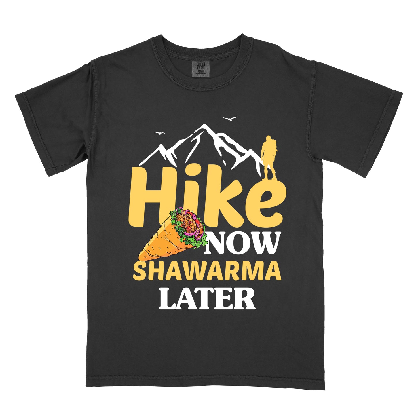 Black t-shirt with text "HIKE NOW SHAWARMA LATER" in yellow and white lettering. A mountain silhouette appears above with a small hiker figure. The word "HIKE" is in large yellow text, "NOW" in smaller white text, "SHAWARMA" in yellow with an illustration of a shawarma wrap beside it, and "LATER" in white. The design emphasizes hiking motivation with food reward.