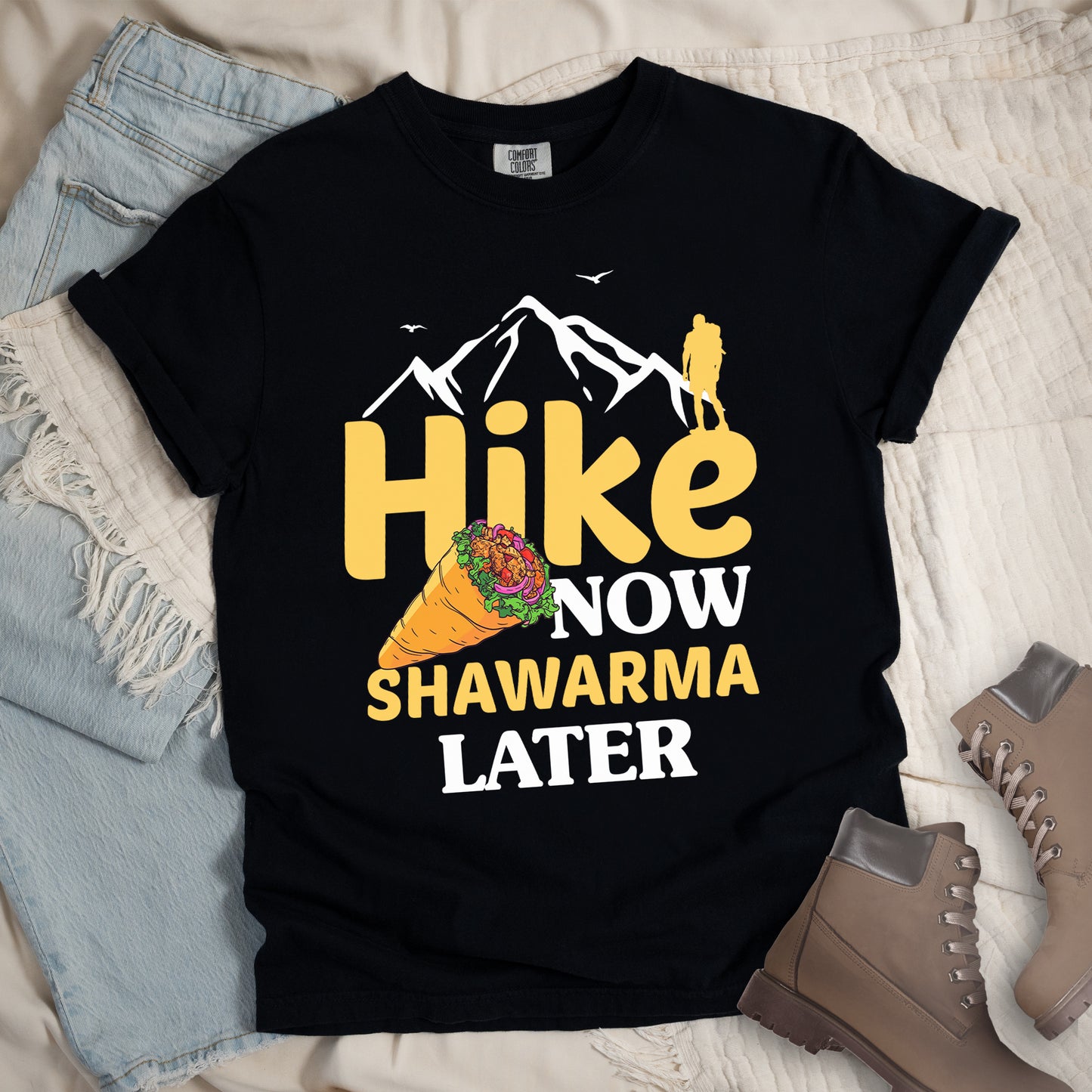 Black t-shirt with text "HIKE NOW SHAWARMA LATER" in yellow and white lettering. A mountain silhouette appears above with a small hiker figure. The word "HIKE" is in large yellow text, "NOW" in smaller white text, "SHAWARMA" in yellow with an illustration of a shawarma wrap beside it, and "LATER" in white. The design emphasizes hiking motivation with food reward. Features a classic crew neck and casual rolled sleeves, styled alongside light wash denim and brown hiking boots.
