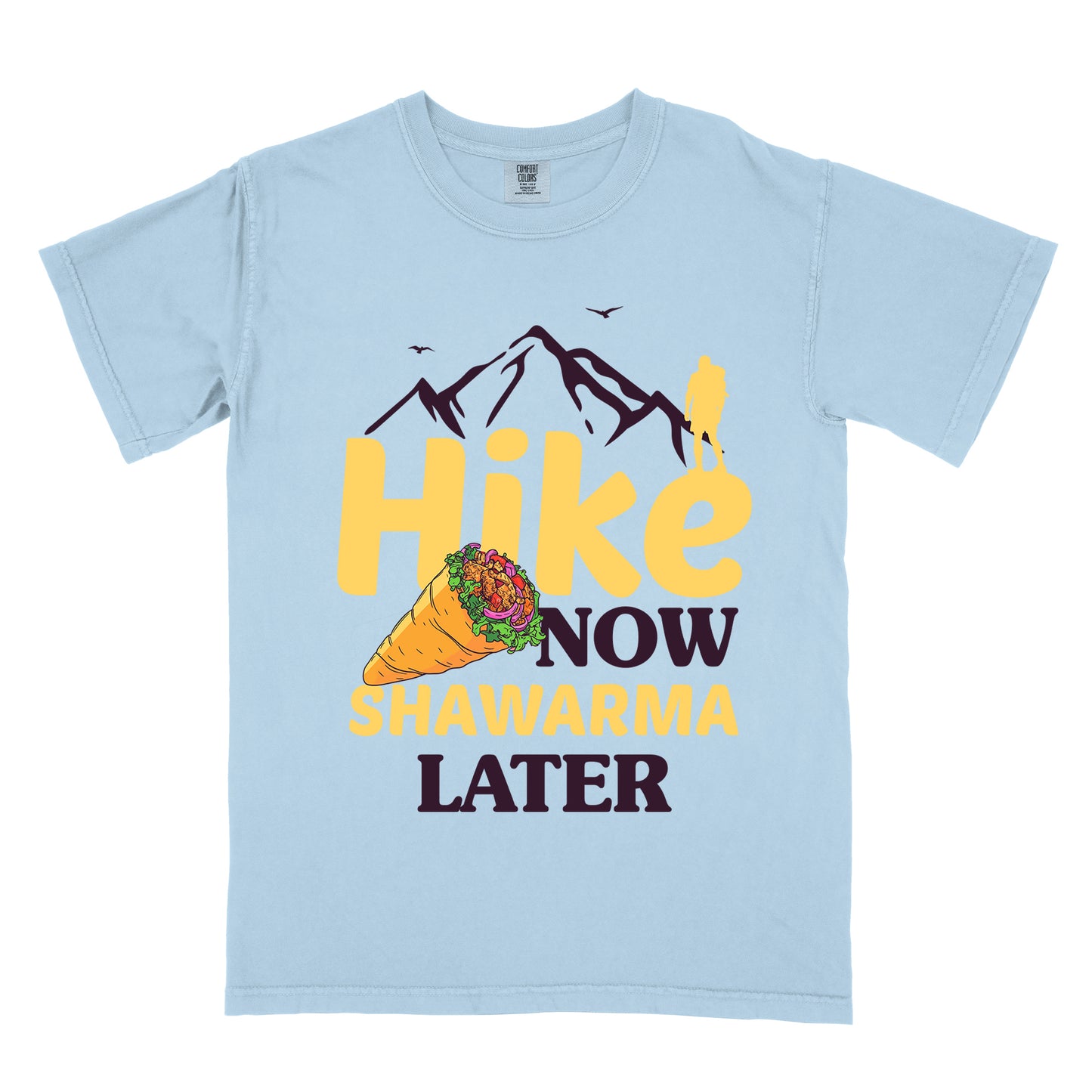 Chambray (Light Blue) t-shirt with text "HIKE NOW SHAWARMA LATER" in yellow and dark brown lettering. A mountain silhouette appears above with a small hiker figure. The word "HIKE" is in large yellow text, "NOW" in smaller dark brown text, "SHAWARMA" in yellow with an illustration of a shawarma wrap beside it, and "LATER" in dark brown. The design emphasizes hiking motivation with food reward.