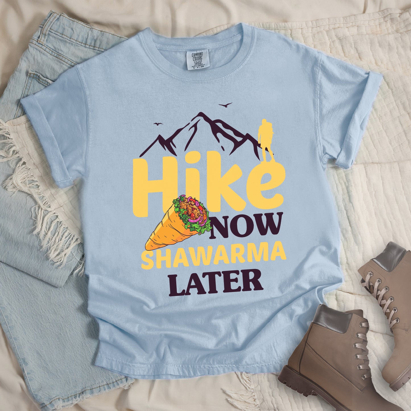 Chambray (Light Blue) t-shirt with text "HIKE NOW SHAWARMA LATER" in yellow and dark brown lettering. A mountain silhouette appears above with a small hiker figure. The word "HIKE" is in large yellow text, "NOW" in smaller dark brown text, "SHAWARMA" in yellow with an illustration of a shawarma wrap beside it, and "LATER" in dark brown. The design emphasizes hiking motivation with food reward. Features a classic crew neck and casual rolled sleeves, styled alongside light wash denim and brown hiking boots.