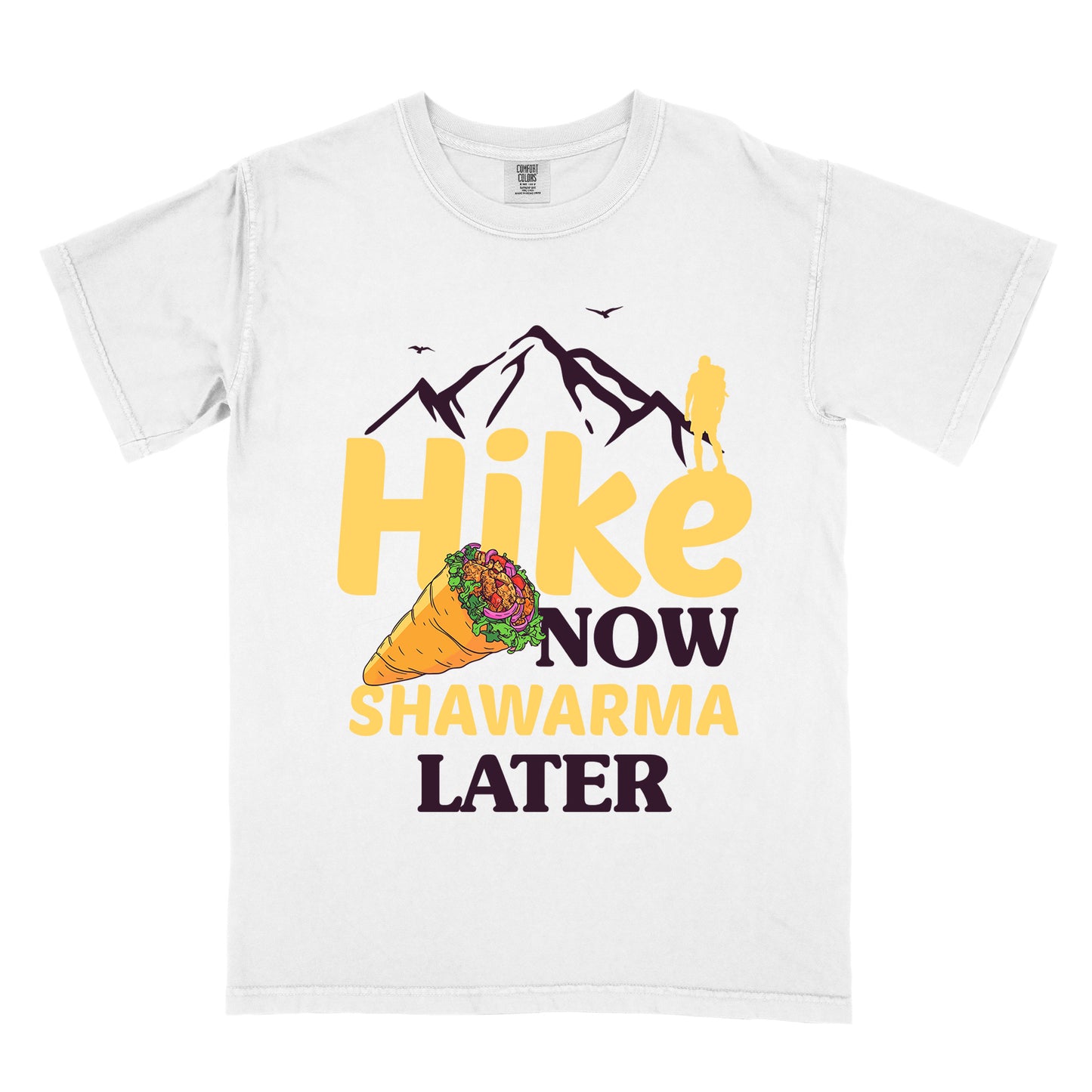 White t-shirt with text "HIKE NOW SHAWARMA LATER" in yellow and dark brown lettering. A mountain silhouette appears above with a small hiker figure. The word "HIKE" is in large yellow text, "NOW" in smaller dark brown text, "SHAWARMA" in yellow with an illustration of a shawarma wrap beside it, and "LATER" in dark brown. The design emphasizes hiking motivation with food reward.