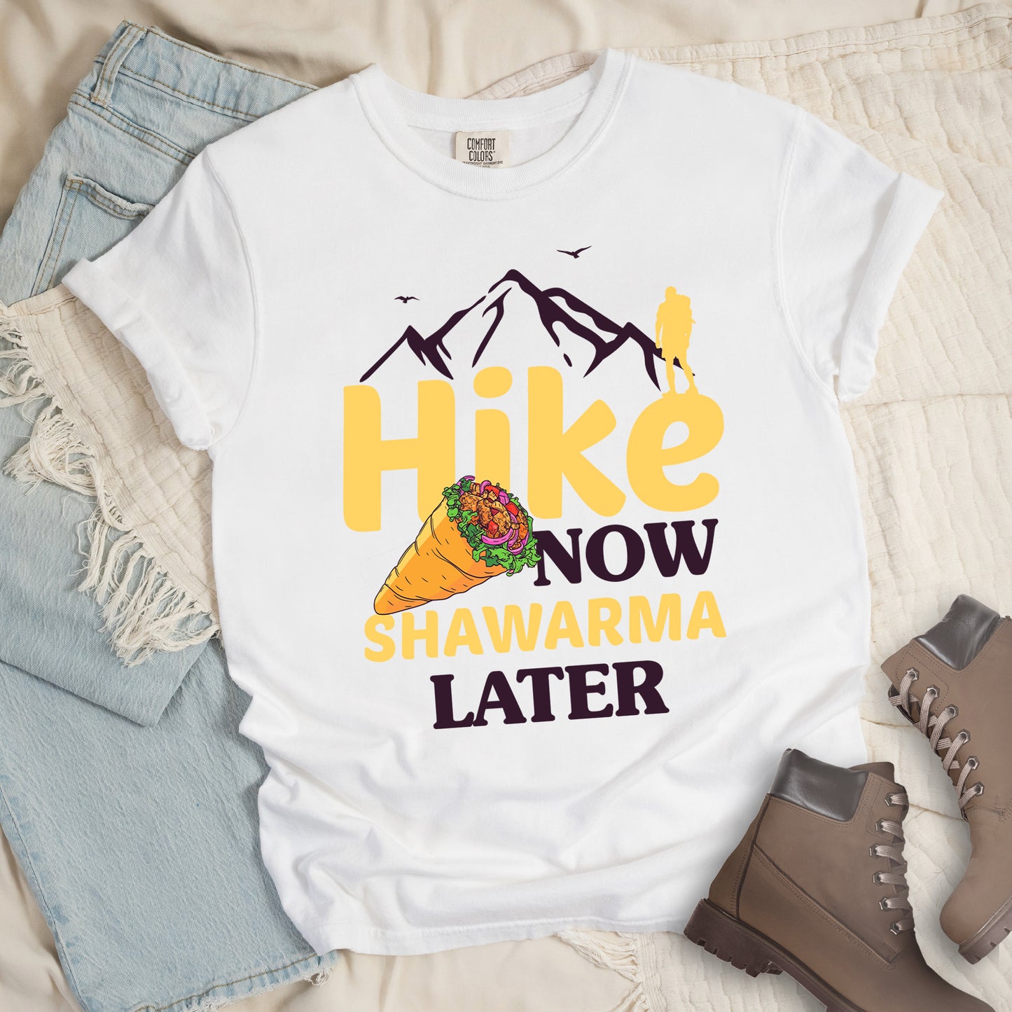 White t-shirt with text "HIKE NOW SHAWARMA LATER" in yellow and dark brown lettering. A mountain silhouette appears above with a small hiker figure. The word "HIKE" is in large yellow text, "NOW" in smaller dark brown text, "SHAWARMA" in yellow with an illustration of a shawarma wrap beside it, and "LATER" in dark brown. The design emphasizes hiking motivation with food reward. Features a classic crew neck and casual rolled sleeves, styled alongside light wash denim and brown hiking boots.